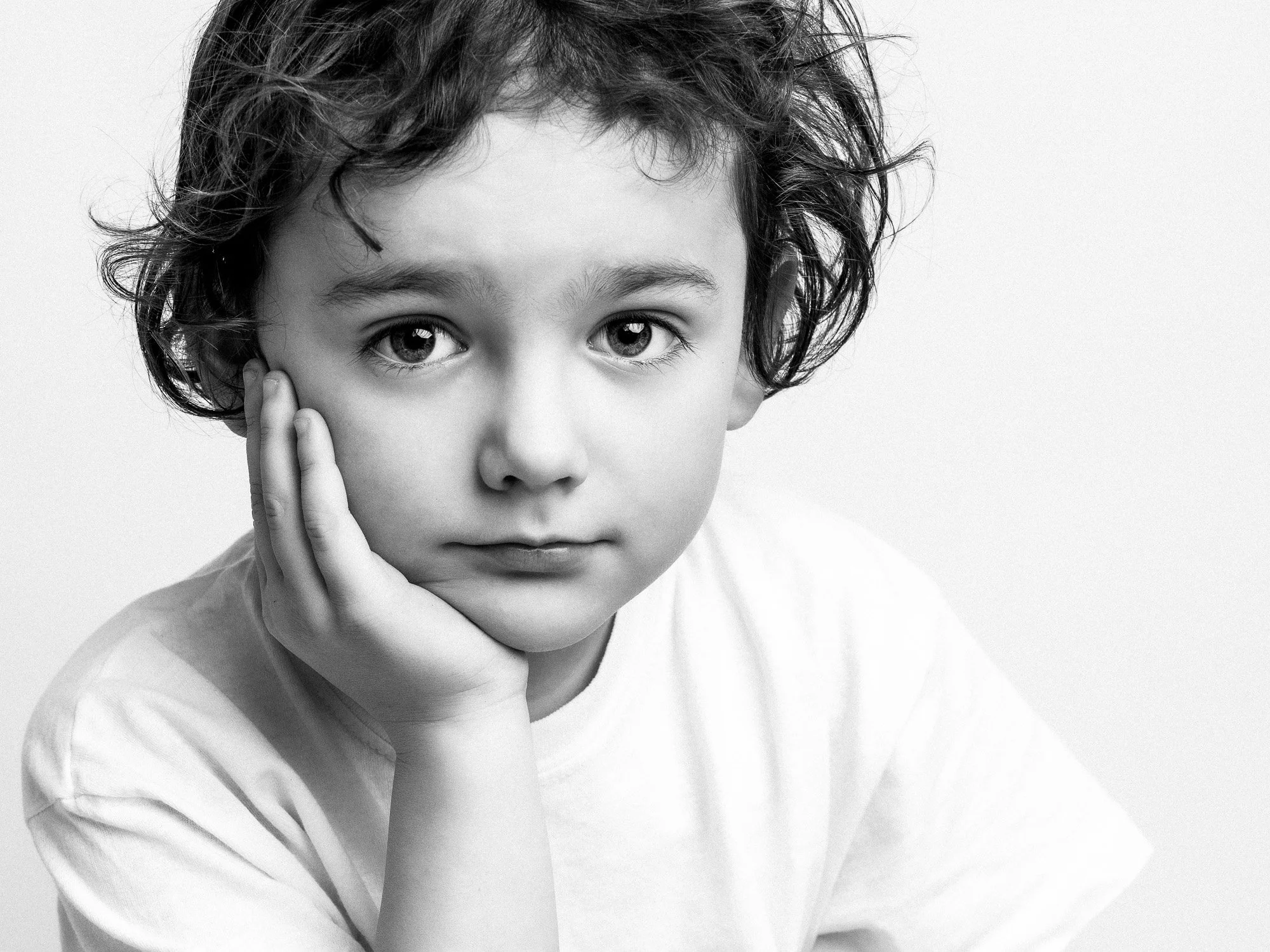 A young child with curly hair resting their head on their hand, looking directly at the camera in black and white.