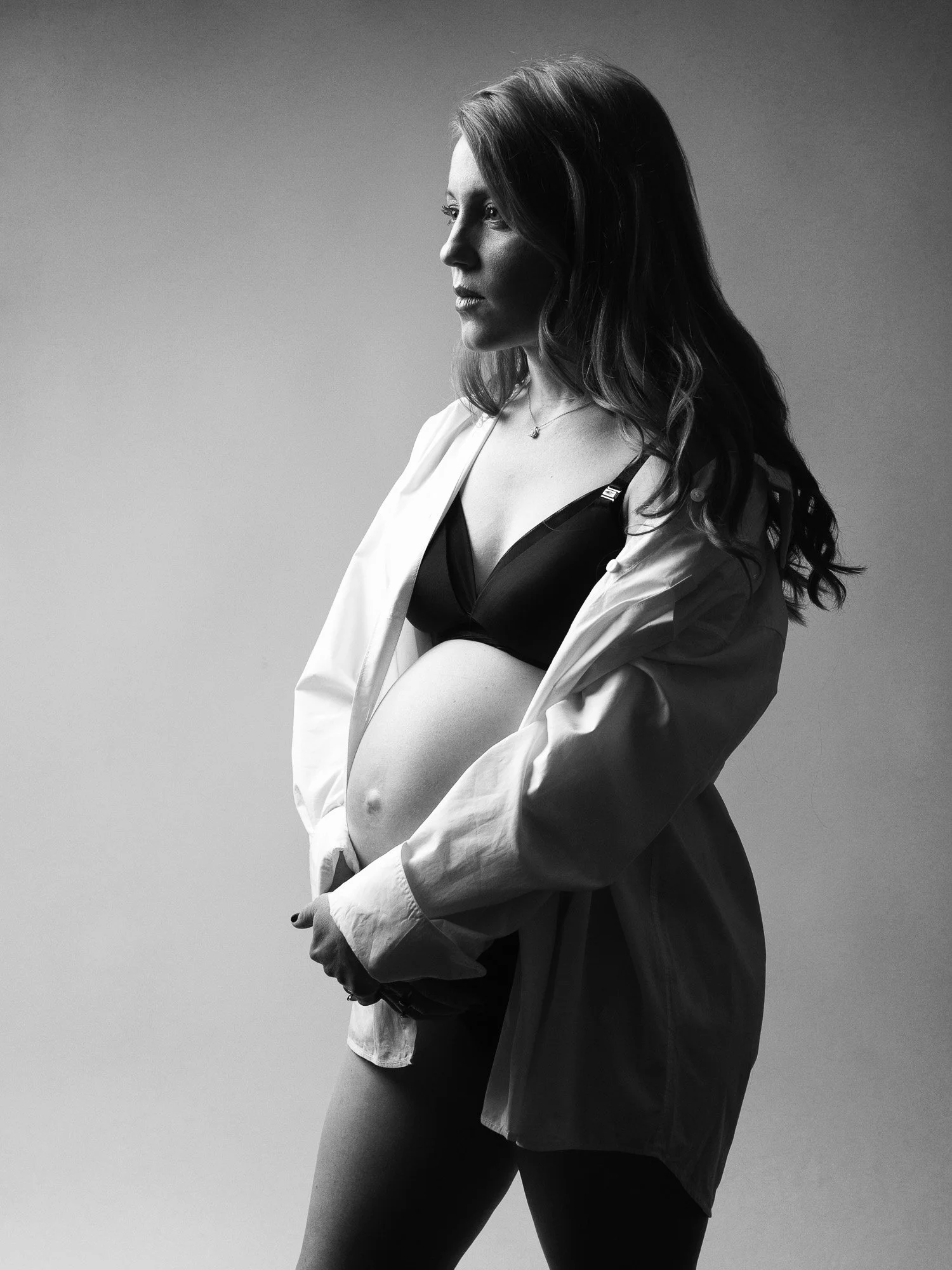 Black and white photo of a pregnant woman looking to the side, wearing a black bra and a loose white shirt.