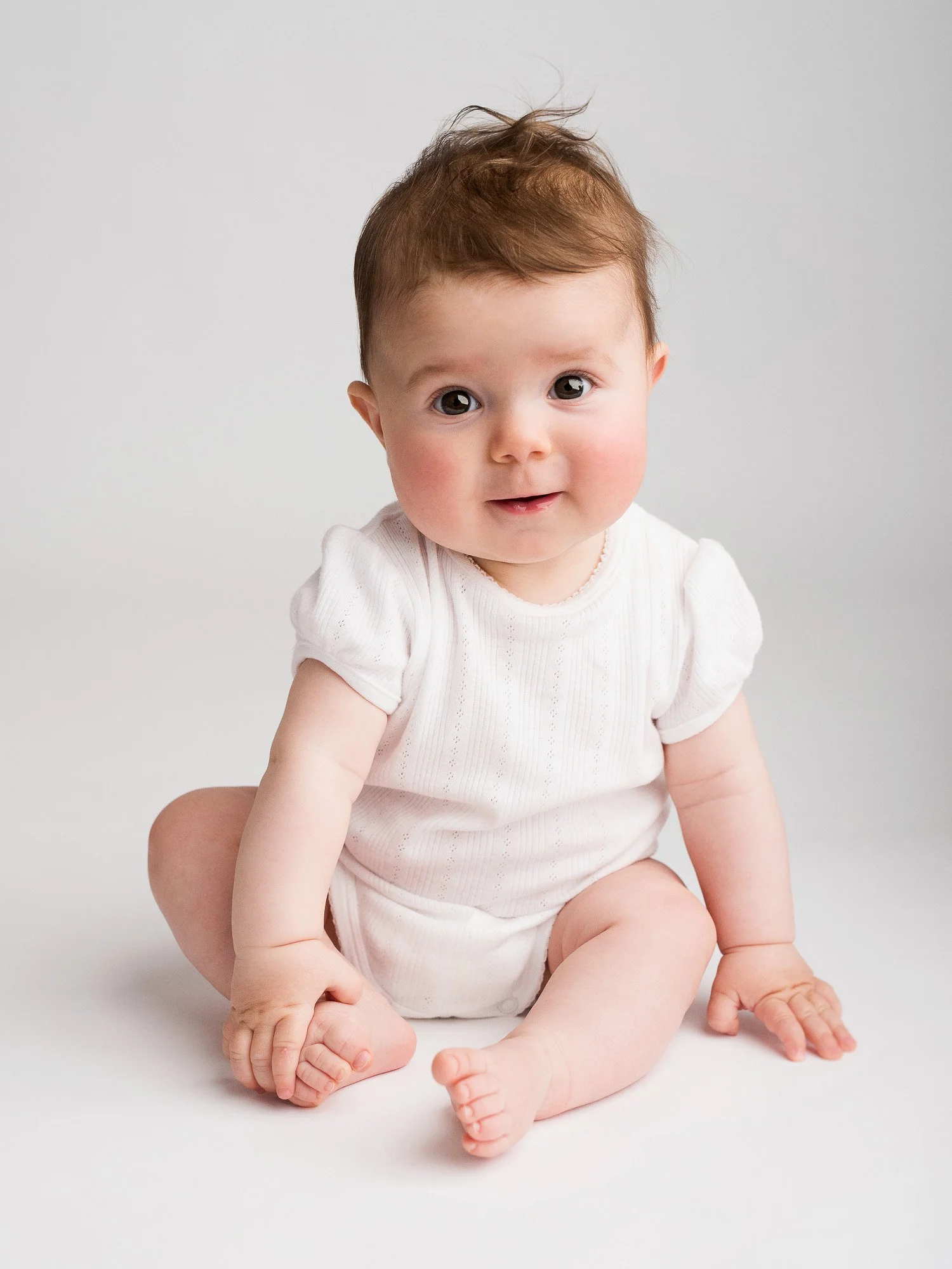 Adorable baby with brown hair sitting on the floor, wearing a white outfit, looking directly at the camera with a neutral expression.