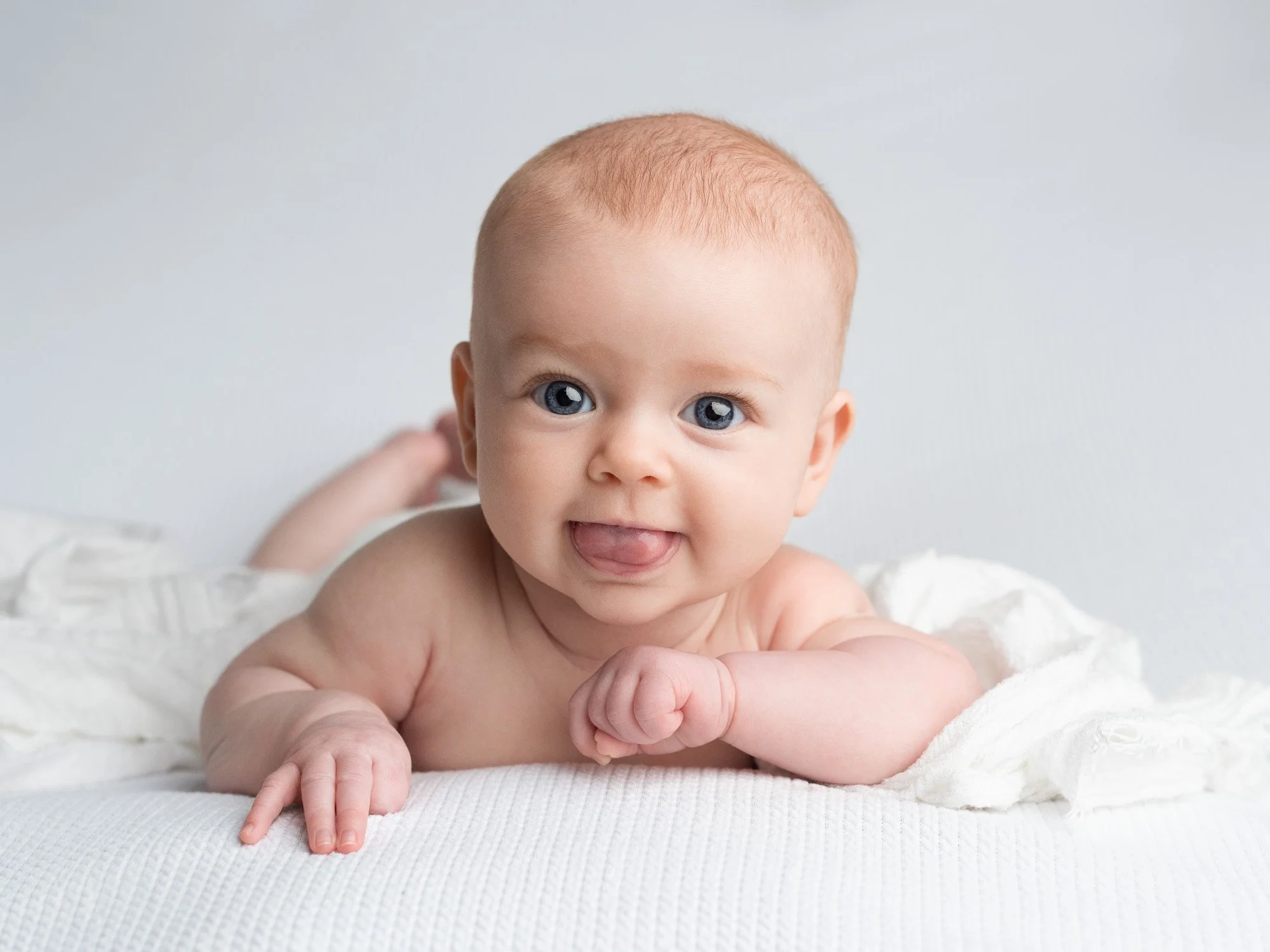 A baby with blue eyes and light red hair lying on a white blanket, looking at the camera with tongue slightly out.