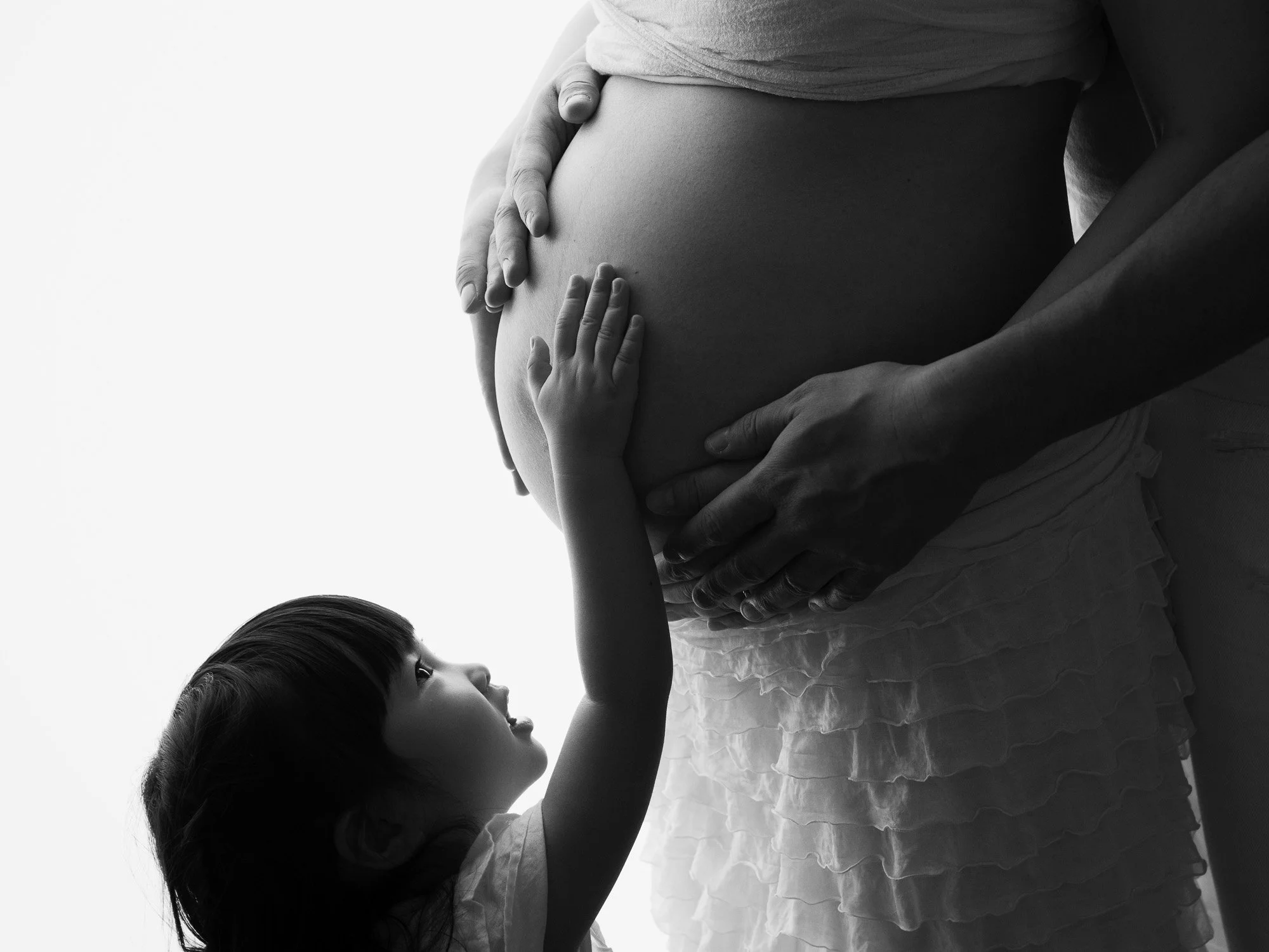 A child gently touching a pregnant woman's belly while looking up at her, with both hands resting on her stomach, black and white photograph.