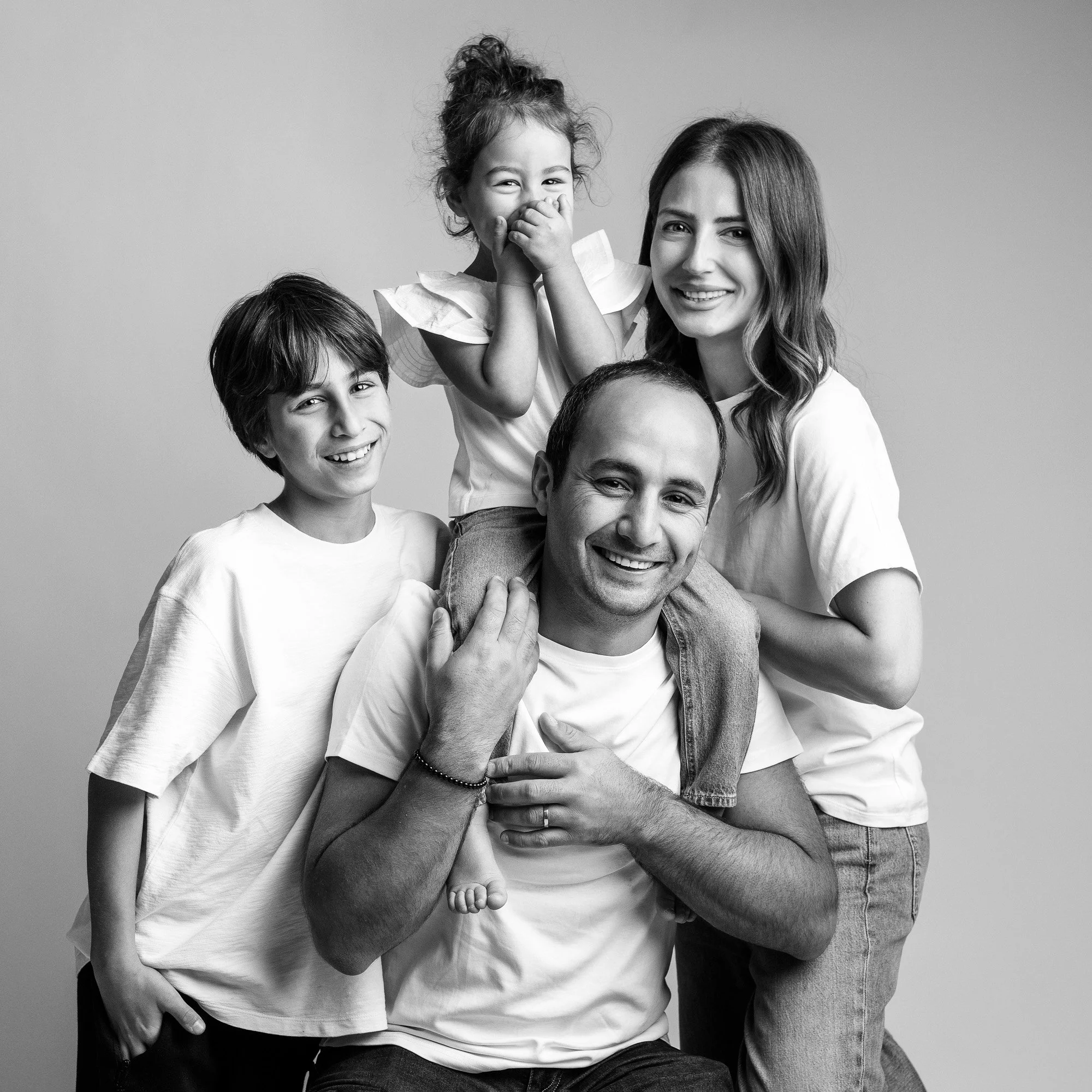 Black and white photo of a family of four smiling, with a father seated and a mother, a boy and a girl standing around him, all wearing casual clothes.