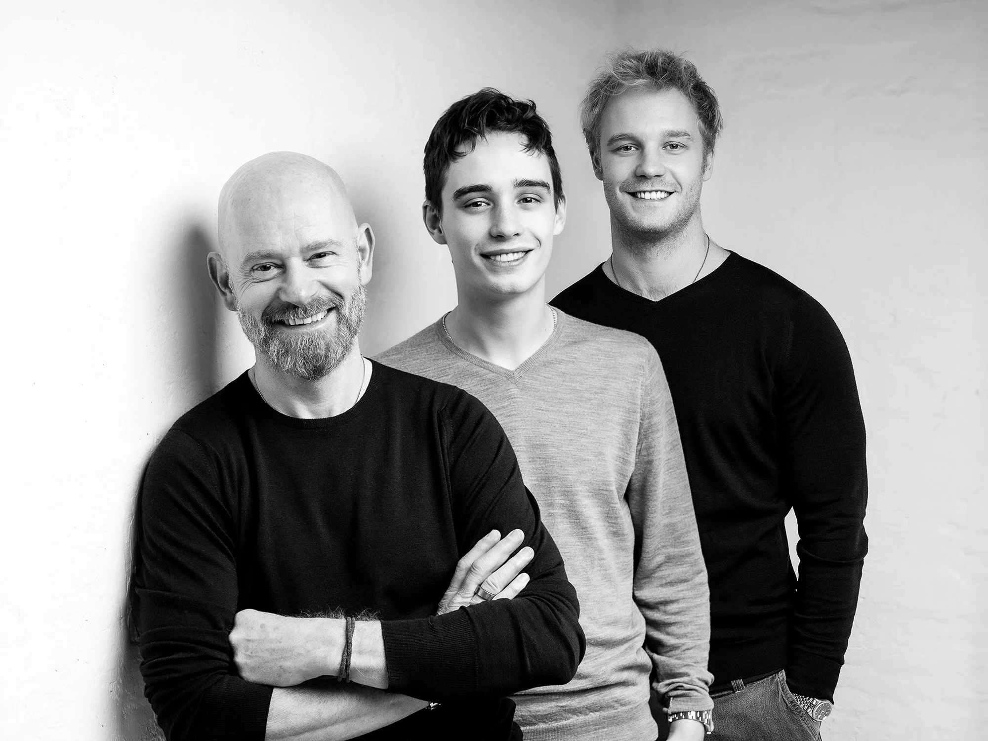 Black and white photo of three men, smiling, standing against a wall. The man on the left has a bald head and a beard, wearing a dark long-sleeve shirt. The man in the middle has short curly hair, wearing a light-colored sweater. The man on the right