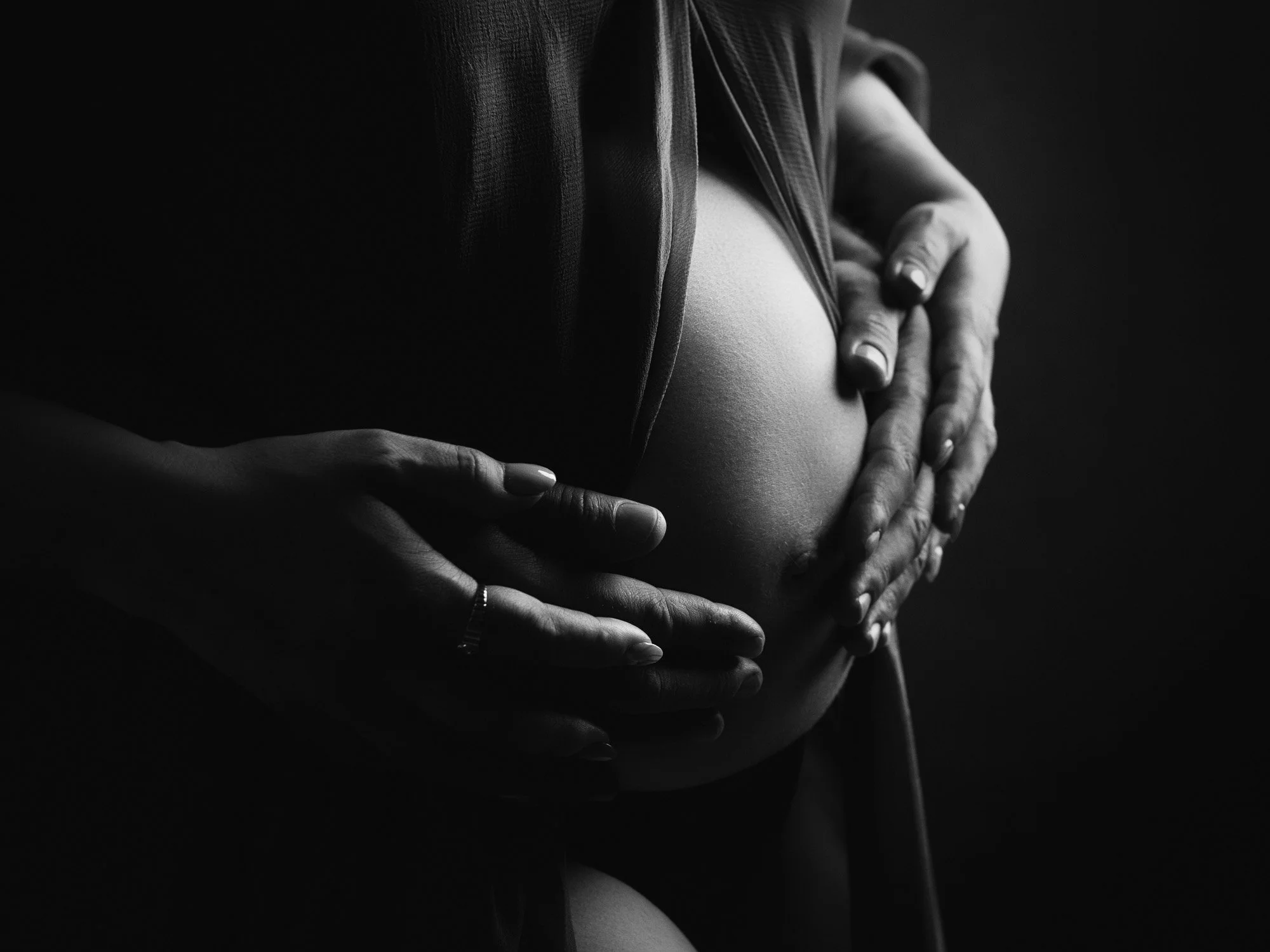Black and white photo of a pregnant woman with her hands gently holding her bare belly, emphasizing the shape of her pregnant stomach.