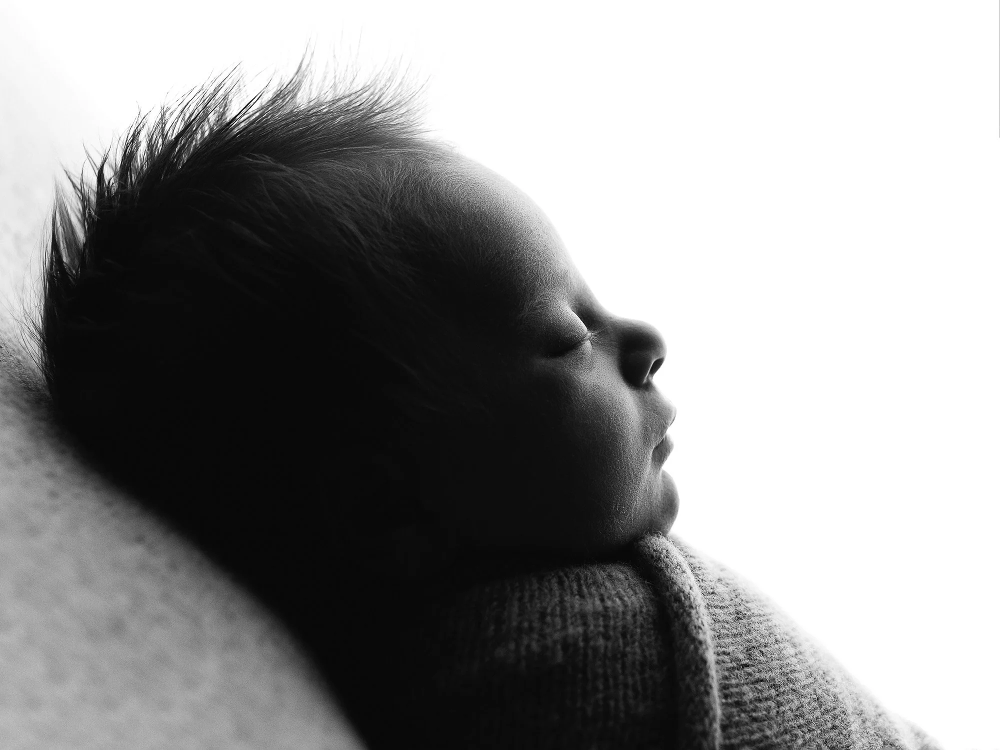 Close-up black and white photo of a newborn baby sleeping peacefully with eyes closed, resting on a soft surface.