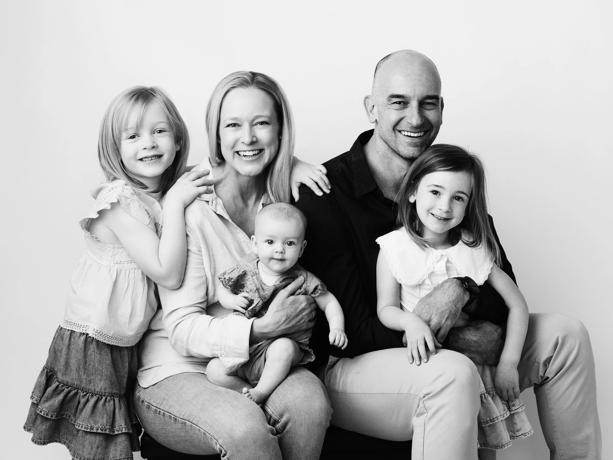 Black and white photo of a smiling family of six, including two adults and four children, sitting close together against a plain background.