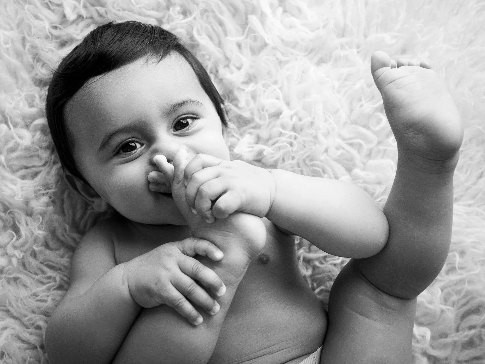 A black and white photo of a smiling baby lying on a fluffy surface, holding their foot and looking at the camera.