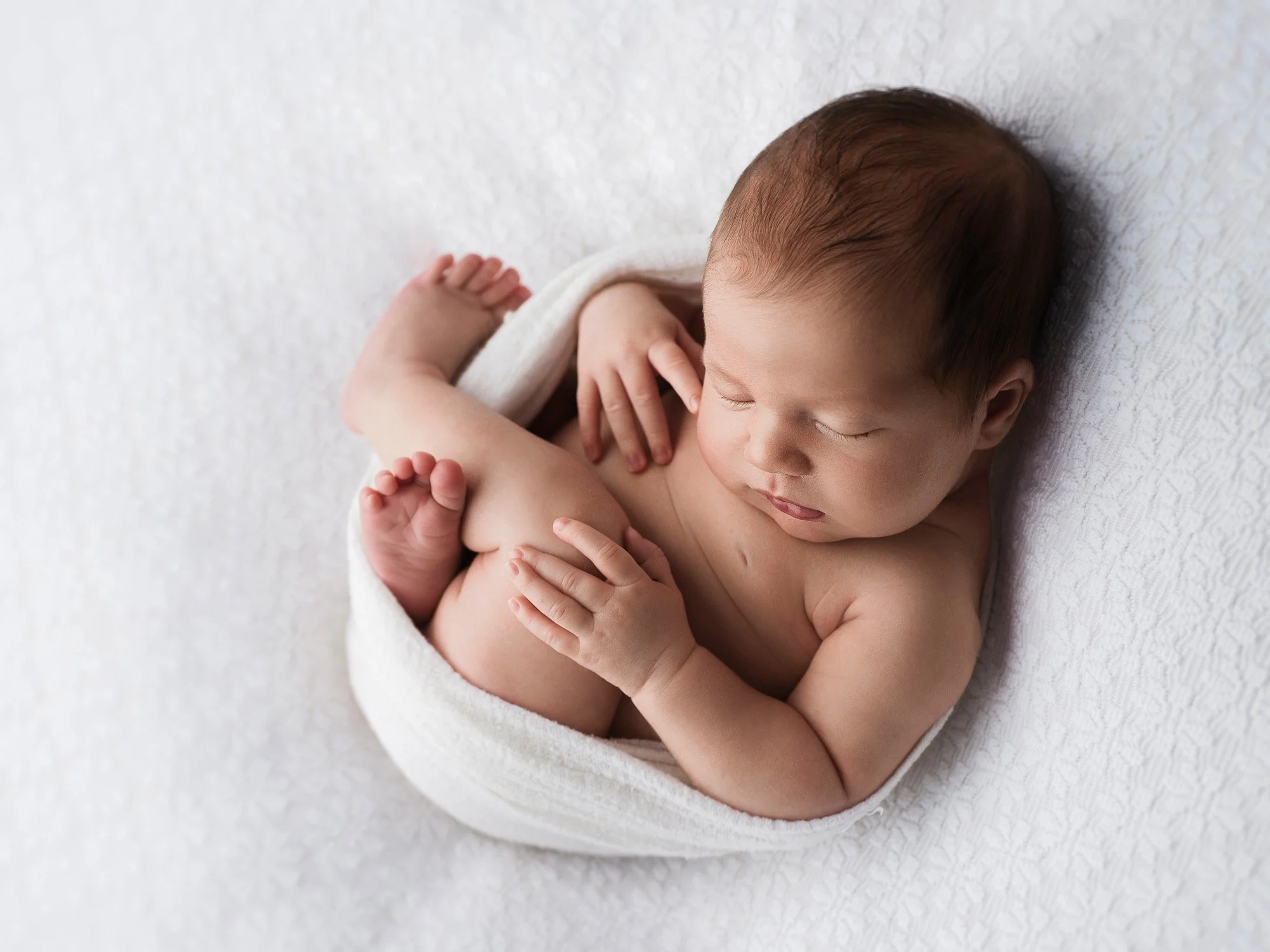 A sleeping baby wrapped in a white blanket on a white textured surface.