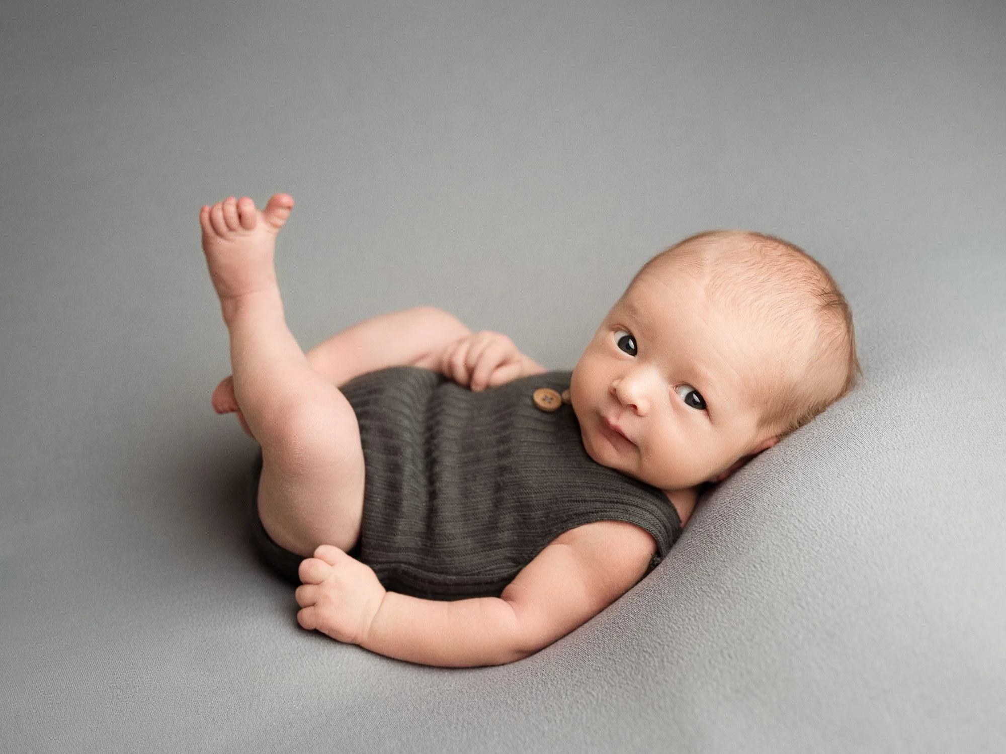 A baby lying on a soft grey surface, wearing a dark grey sleeveless outfit with a small wooden button, looking directly at the camera.