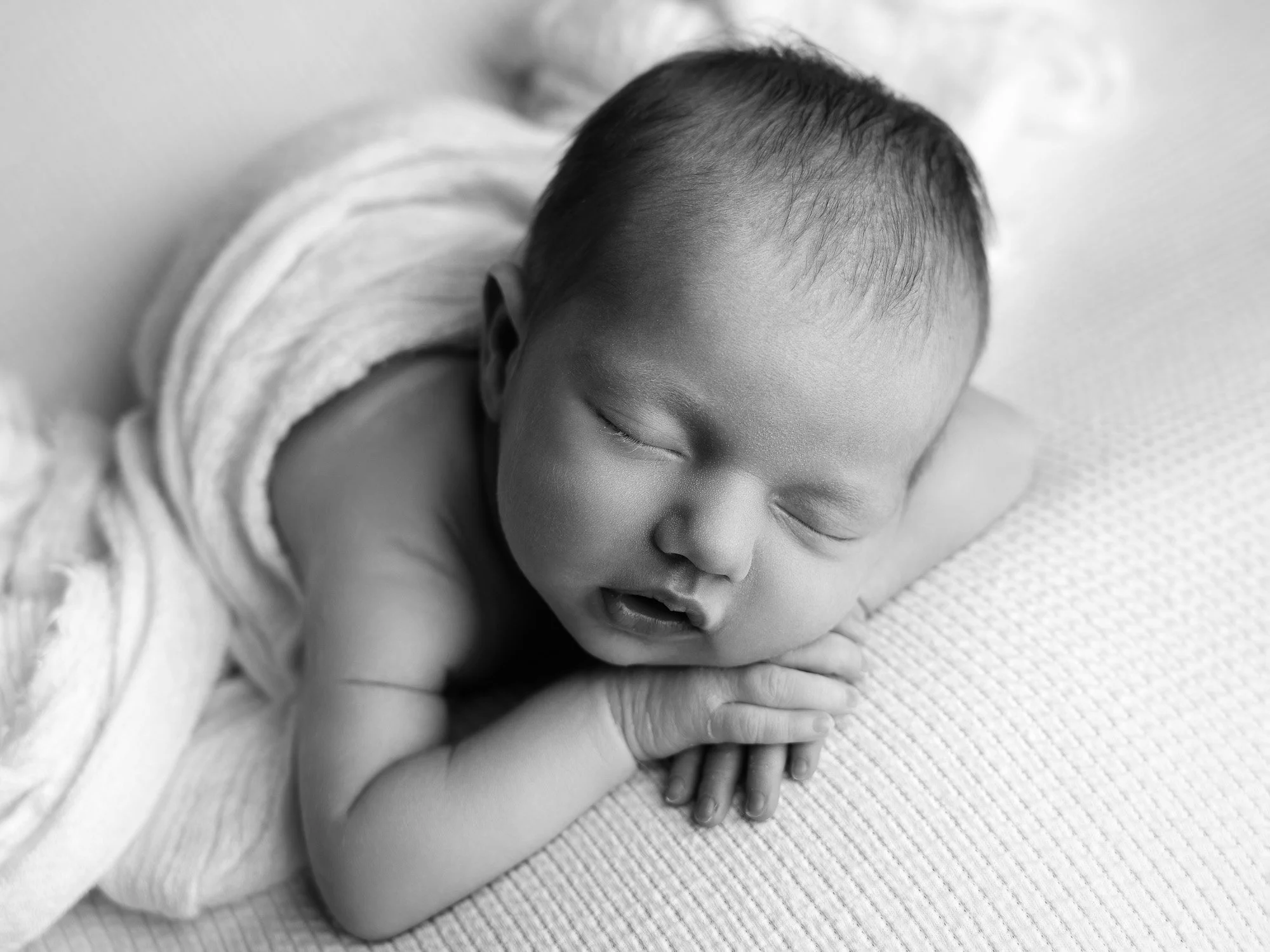 Black and white photo of a sleeping baby resting on a soft surface with hands under the chin and eyes closed.