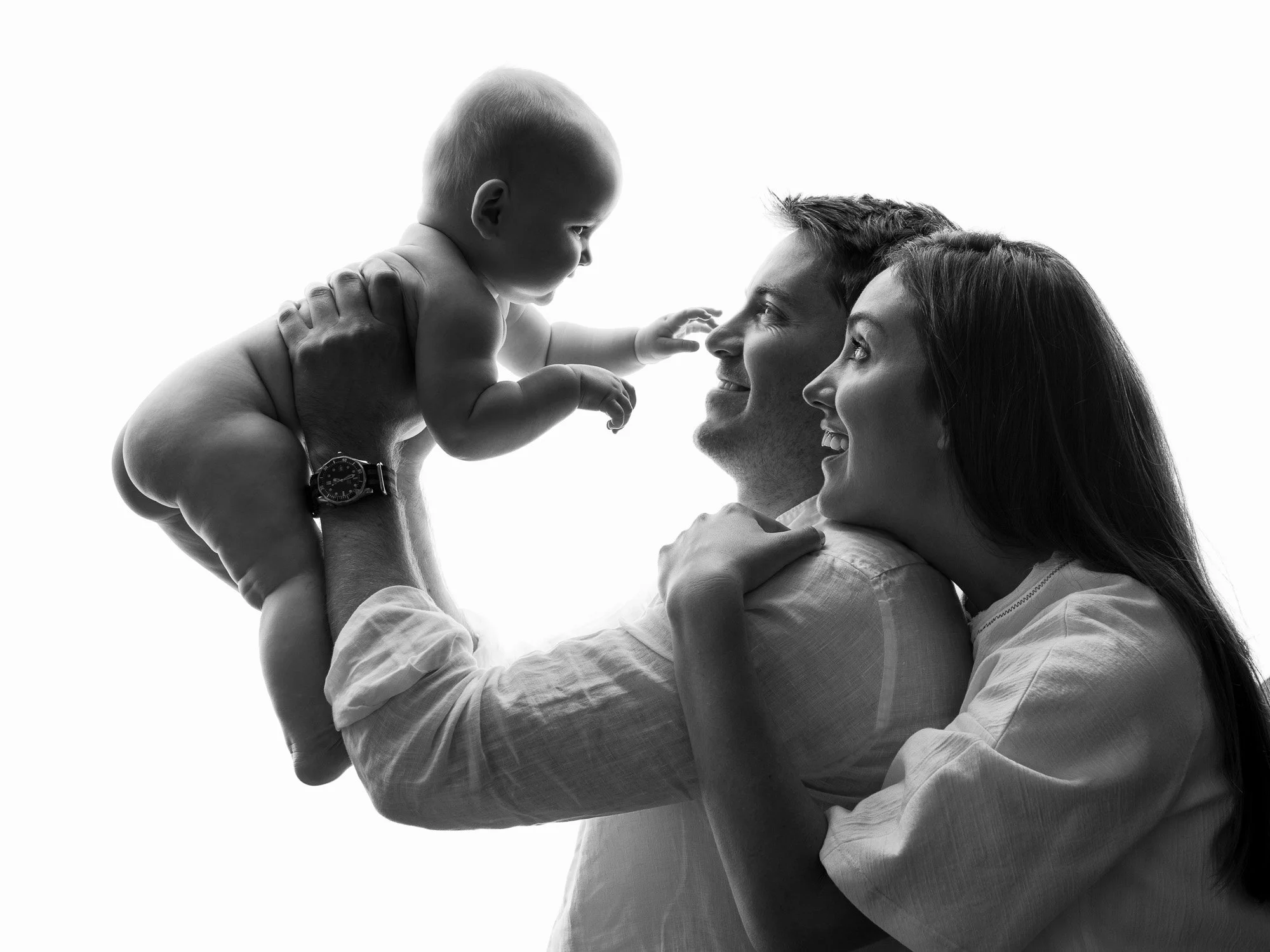 A black-and-white photo of a happy family with a father, mother, and baby. The father is holding the baby in the air, and the baby is reaching out towards the father and mother. The family is smiling and looking at each other, with a white background