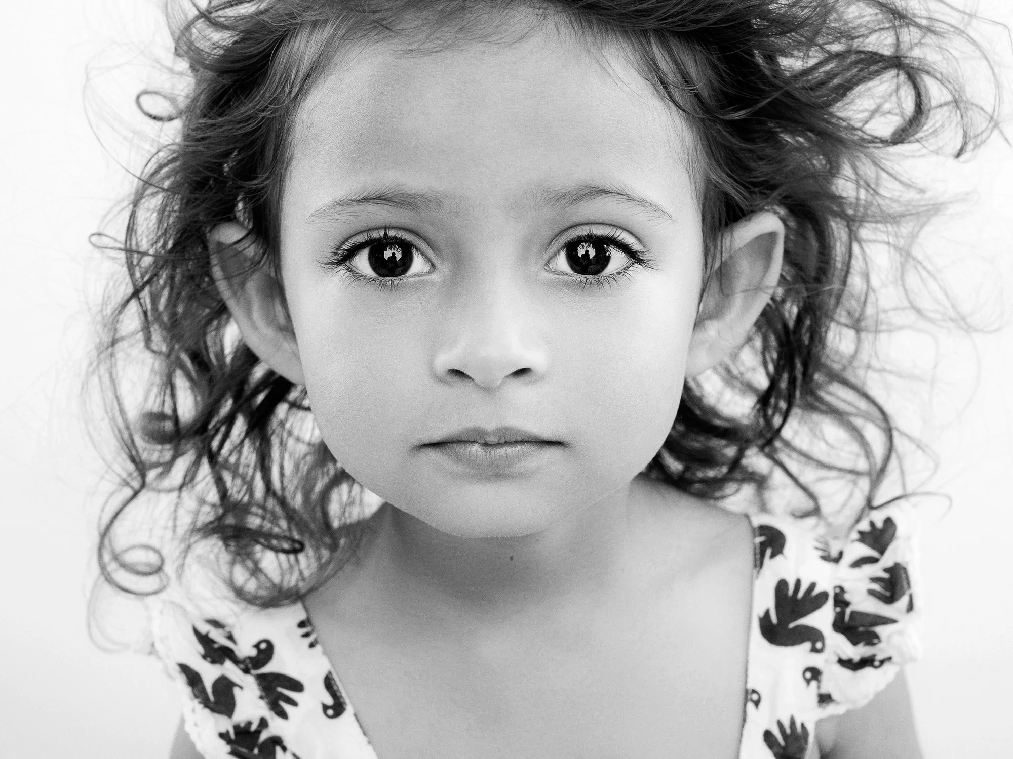 Close-up black and white portrait of a young girl with big eyes, curly hair, and wearing a dress with a handprint pattern, staring directly at the camera.