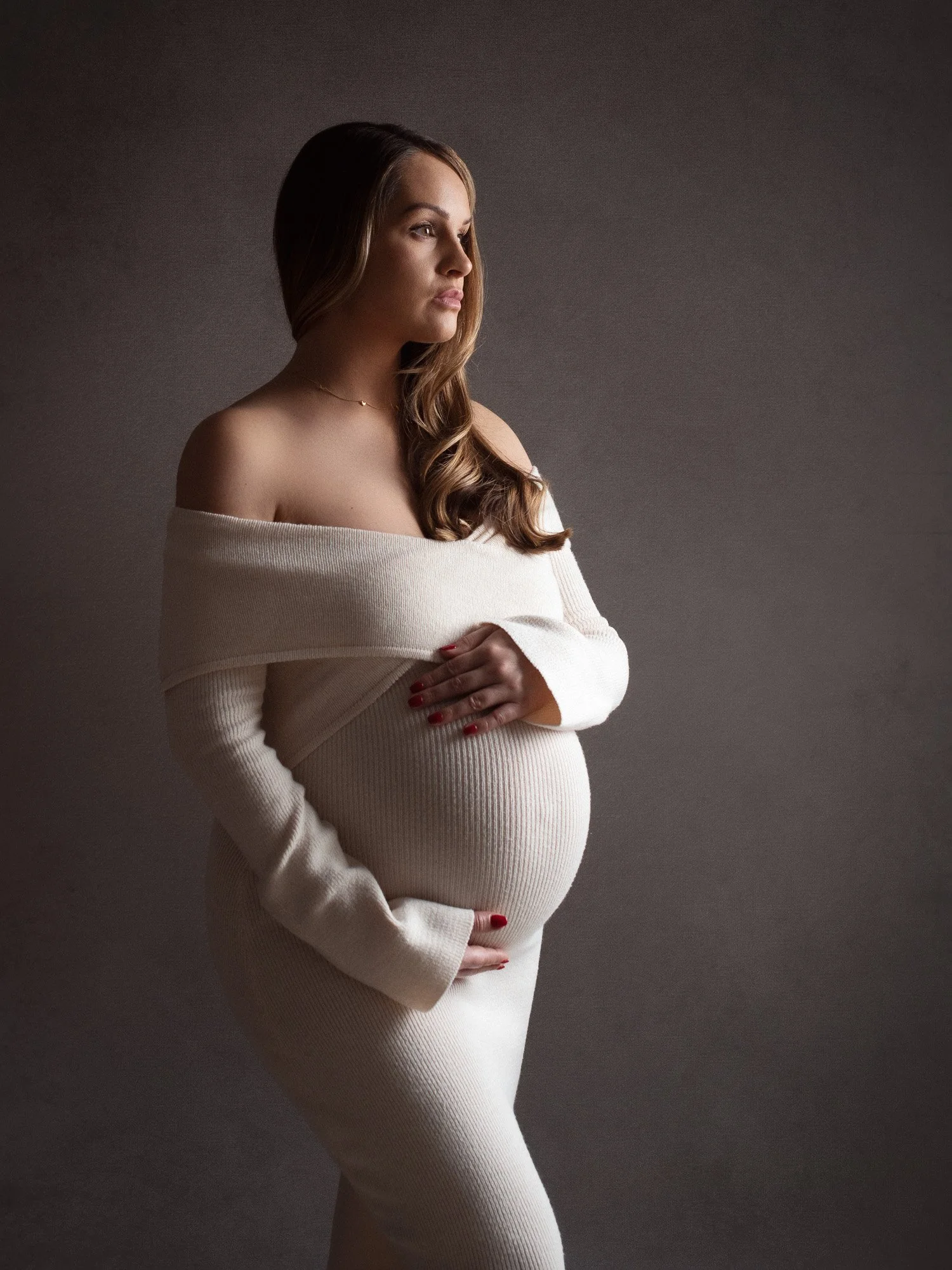 A pregnant woman with long brown hair looking to the side, wearing a cream-colored off-the-shoulder sweater and matching pants, standing against a dark, textured background.