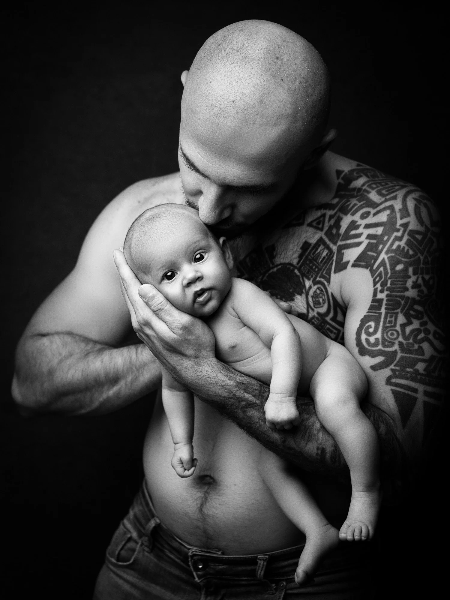 Black and white photo of a bald man with tattoos holding a baby close to his chest, the man is kissing the baby's head, both looking at the camera.