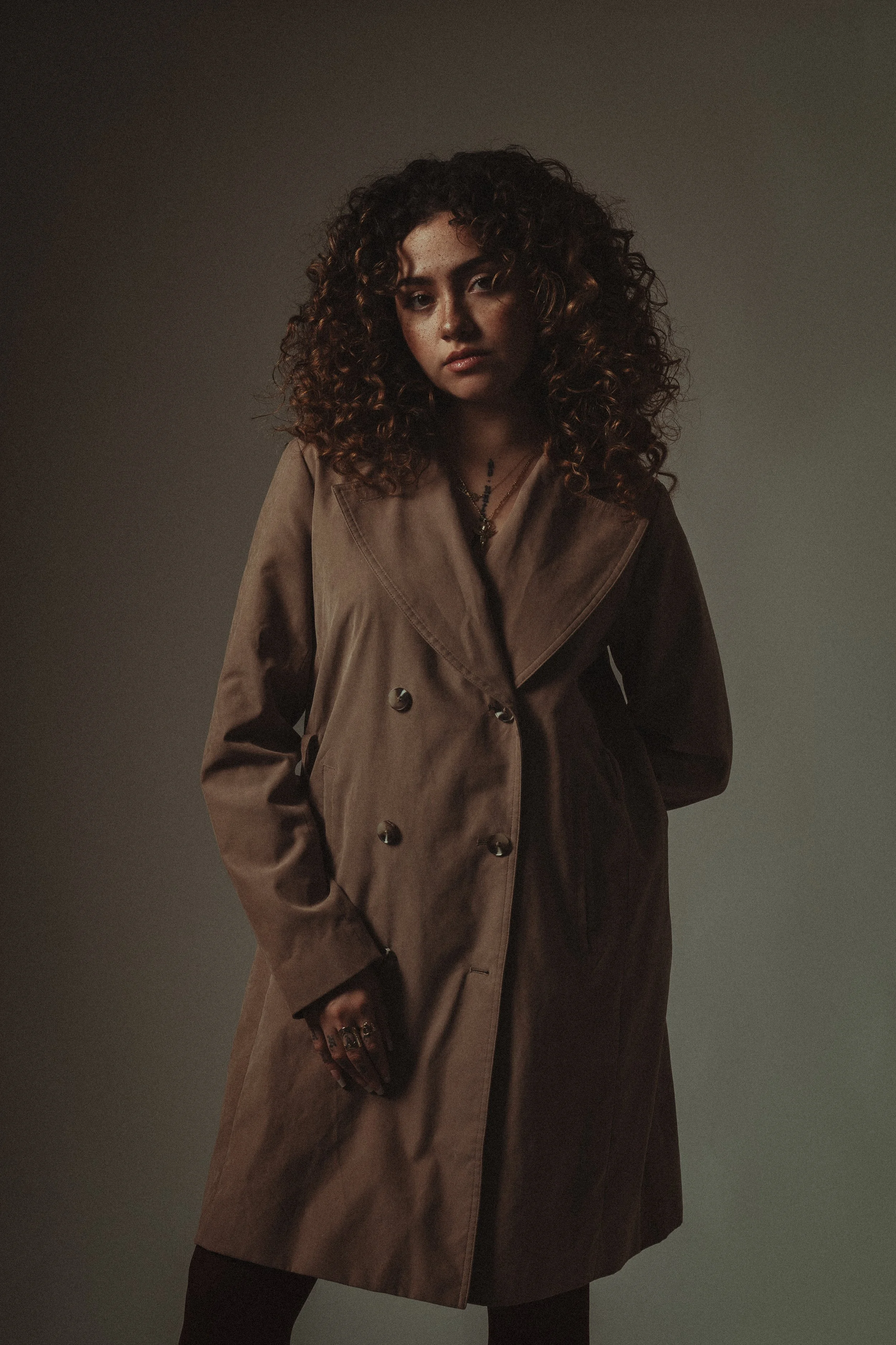Stylish portrait of a woman with curly hair in a brown trench coat, exuding confidence with moody lighting.