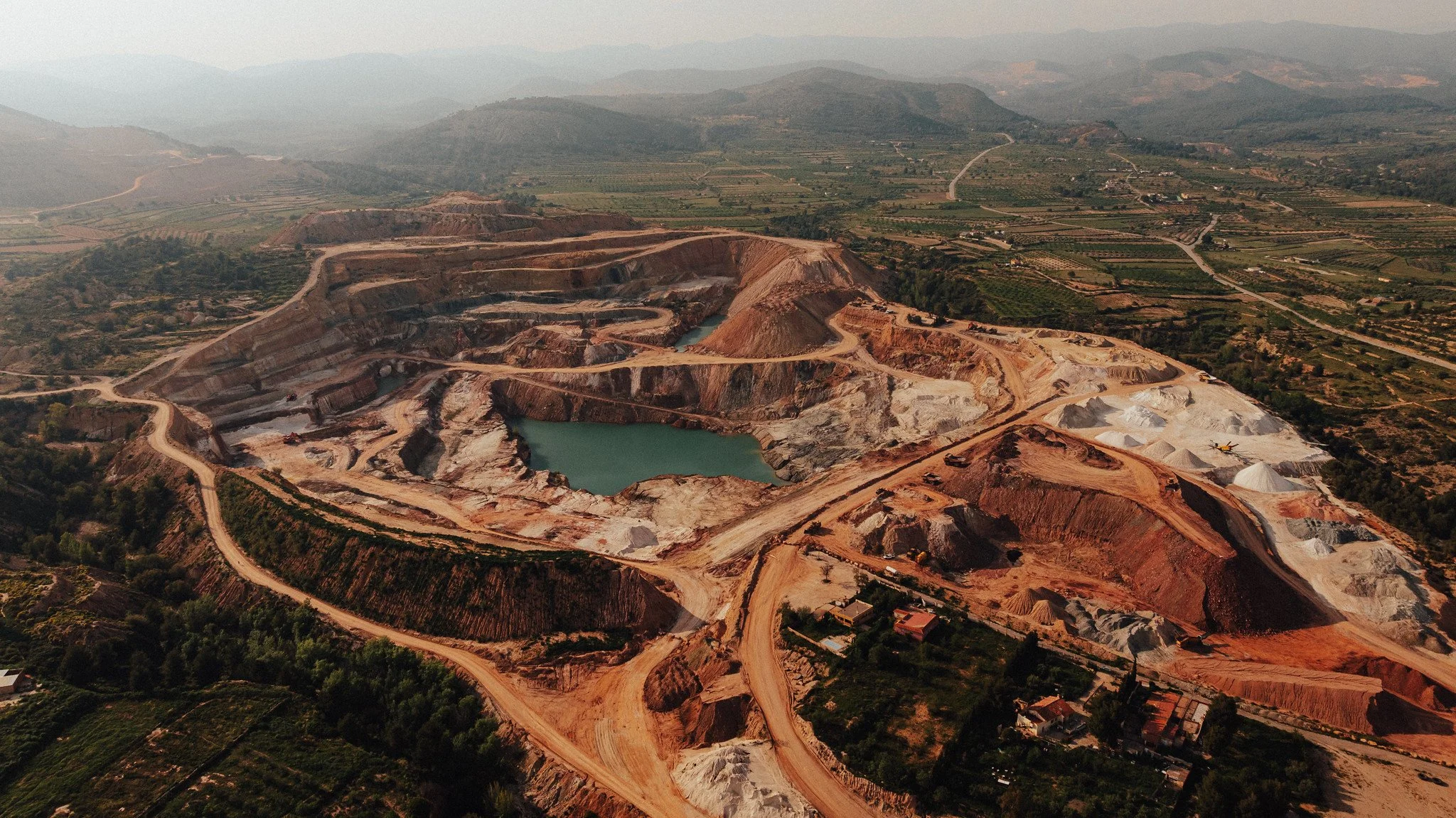 Vista aérea de una cantera con varios estanques, caminos y pilas de materiales, rodeada de tierras de cultivo y montañas al fondo.