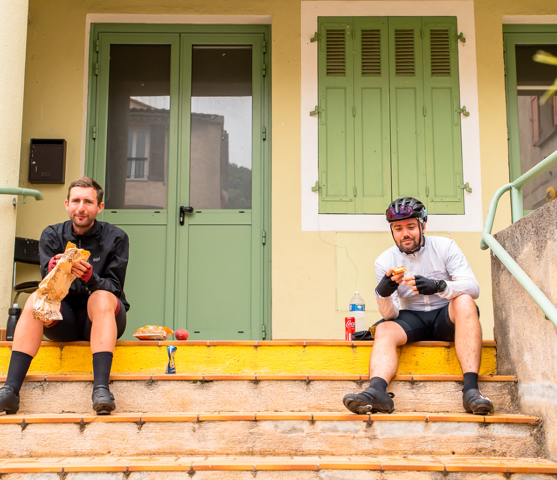 Dos participantes en la Carrera a través de Francia se toman un descanso para comer en las escaleras de un pueblo, ilustrando la sencillez y la resistencia del ultraciclismo.