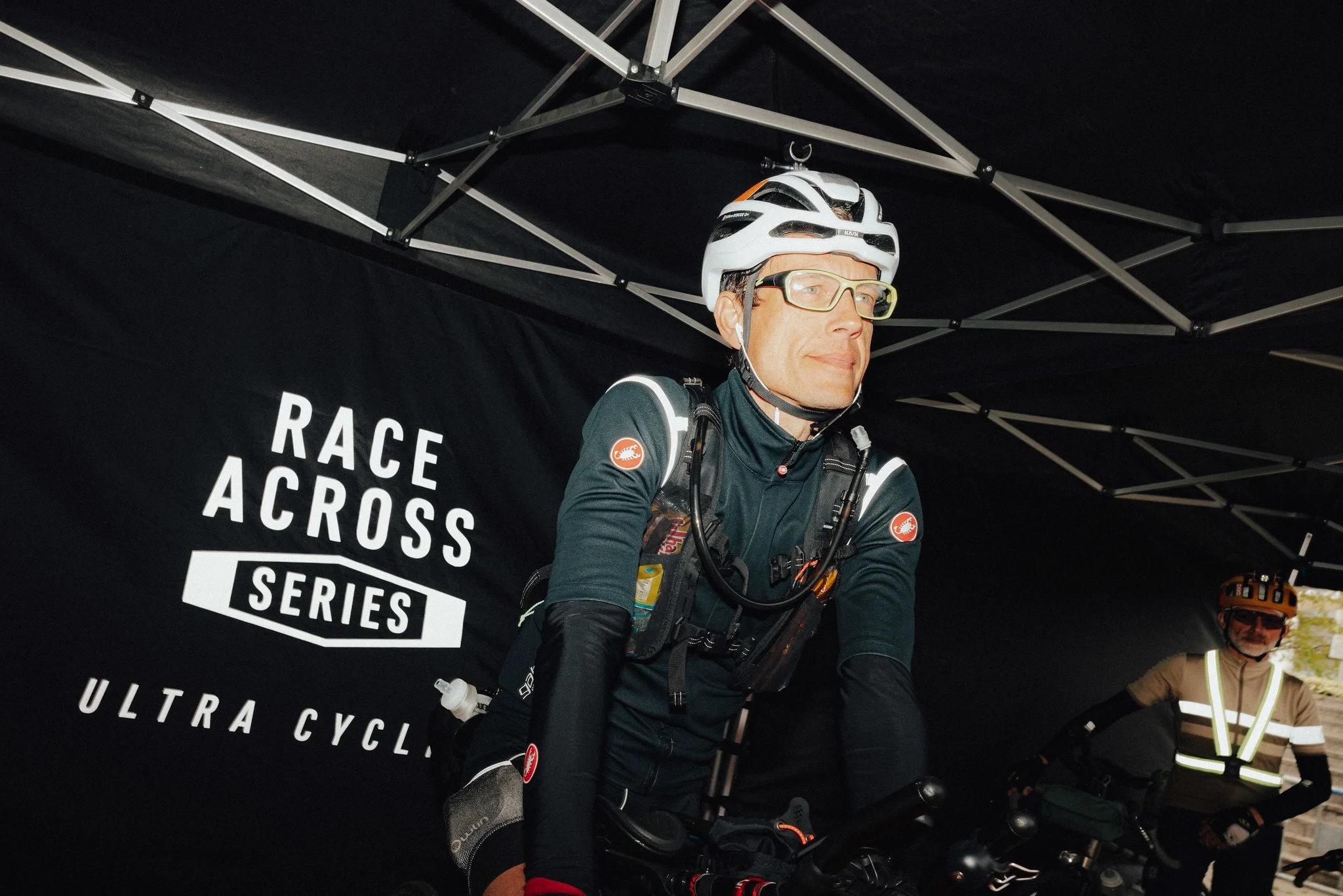Dos ciclistas con equipo de protección, uno de ellos con casco blanco y gafas, se preparan para una carrera de bicicleta de montaña bajo una marquesina negra con el logotipo "Race Across Series Ultra Cyclo".