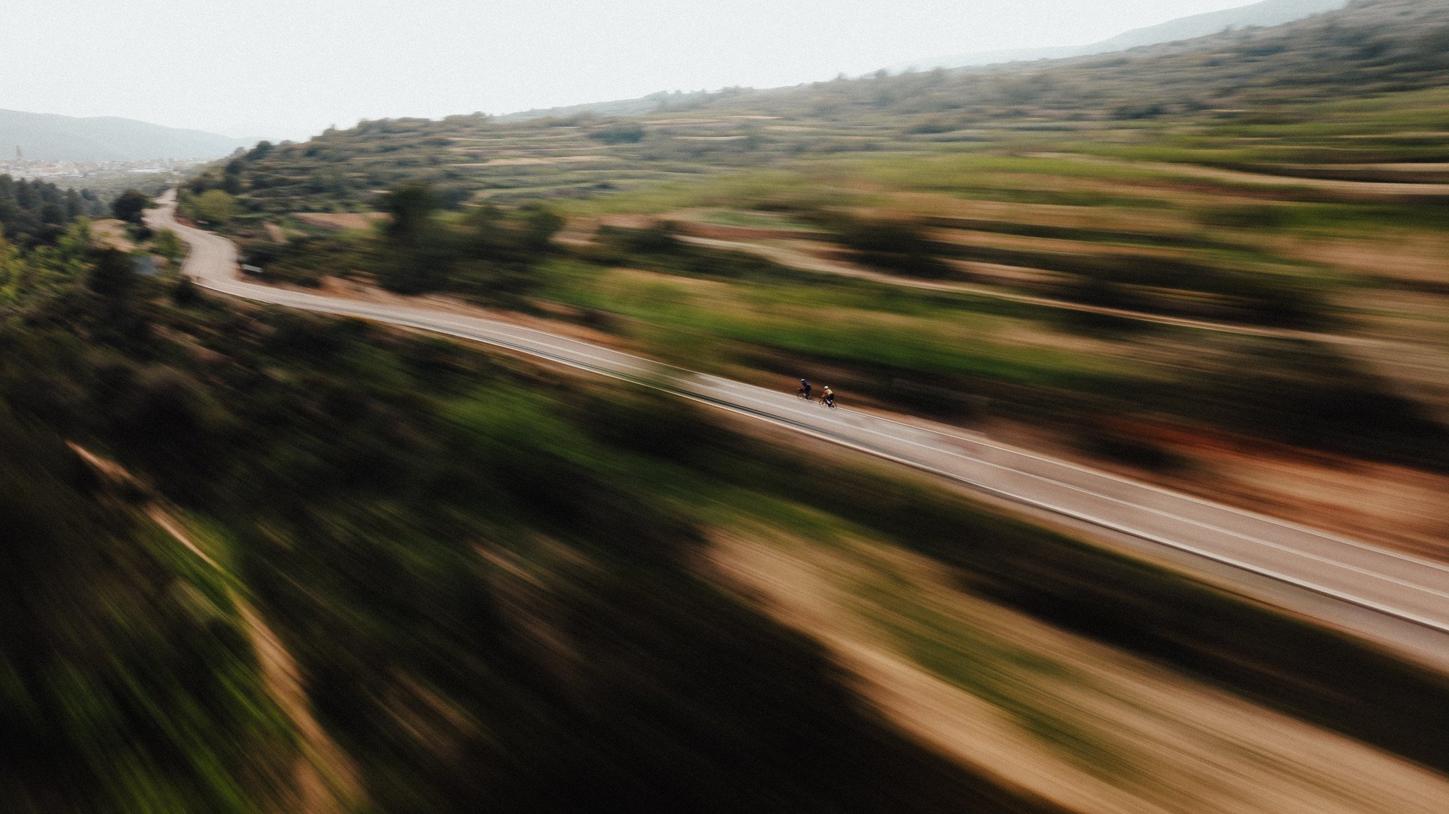 Una carretera serpenteante a través de un paisaje montañoso con dos ciclistas circulando por ella, efectos de movimiento borroso en los alrededores.