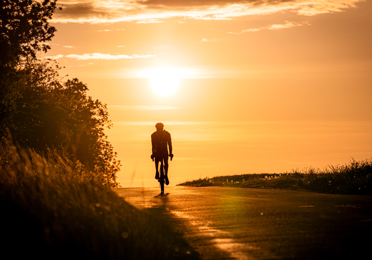 Un cycliste en silhouette roulant au coucher du soleil dans un paysage rural avec champs et arbres à l'arrière-plan.