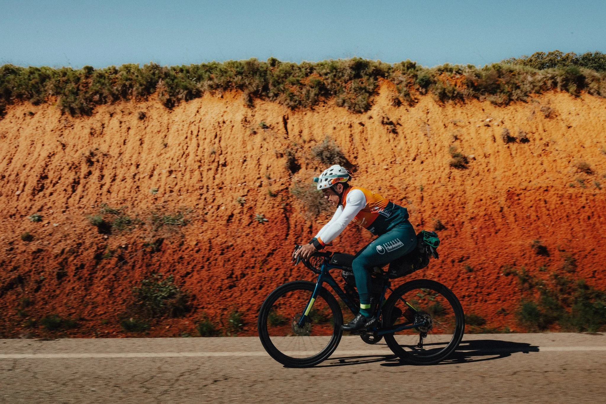 Un ciclista con casco y atuendo colorido pedalea por una carretera con un acantilado rojo de fondo.