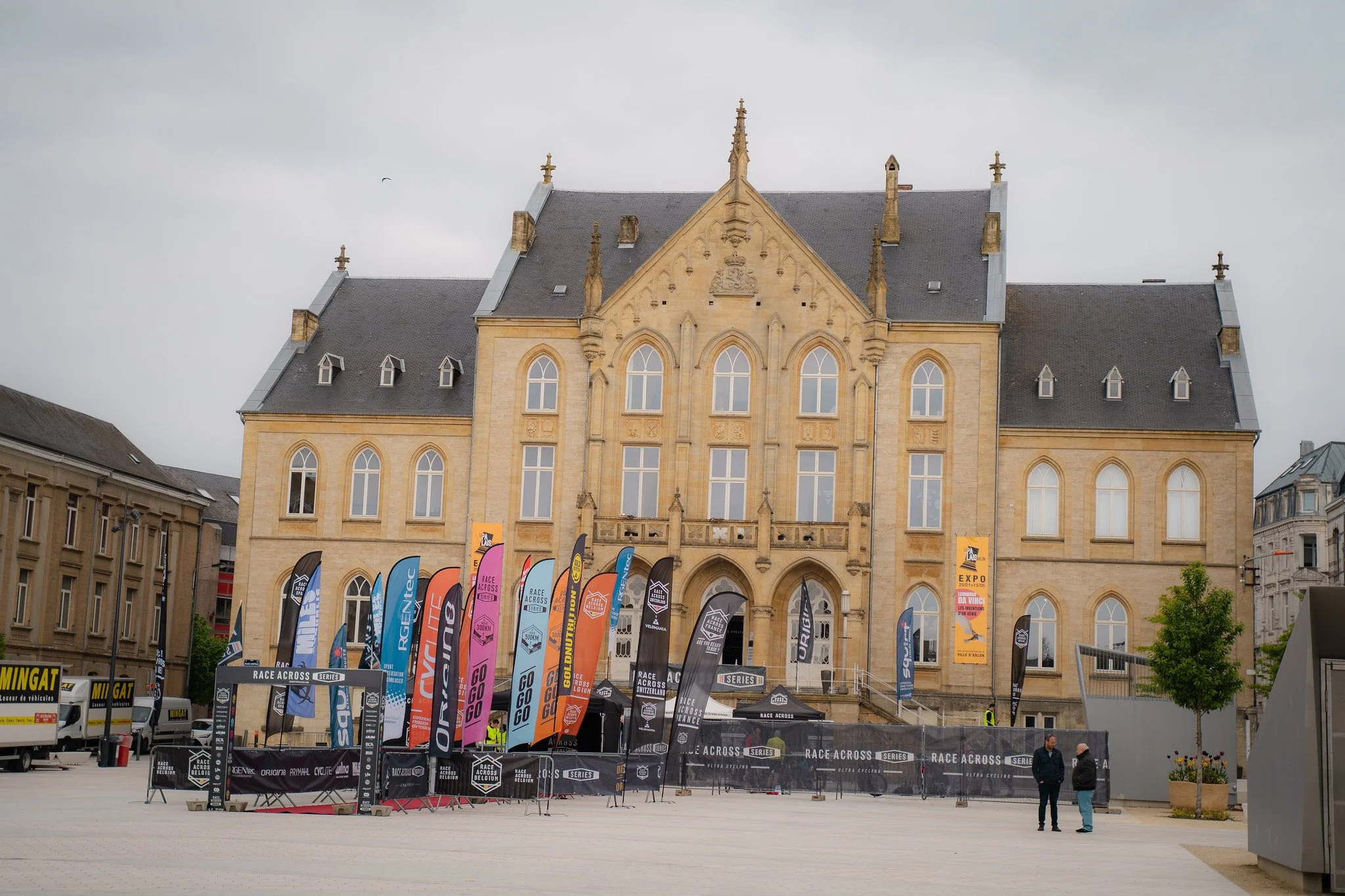 Une place dans une ville avec un bâtiment gothique en pierre beige en arrière-plan et plusieurs drapeaux colorés pour un événement de course dans la première ligne. Deux personnes discutent près de la scène.