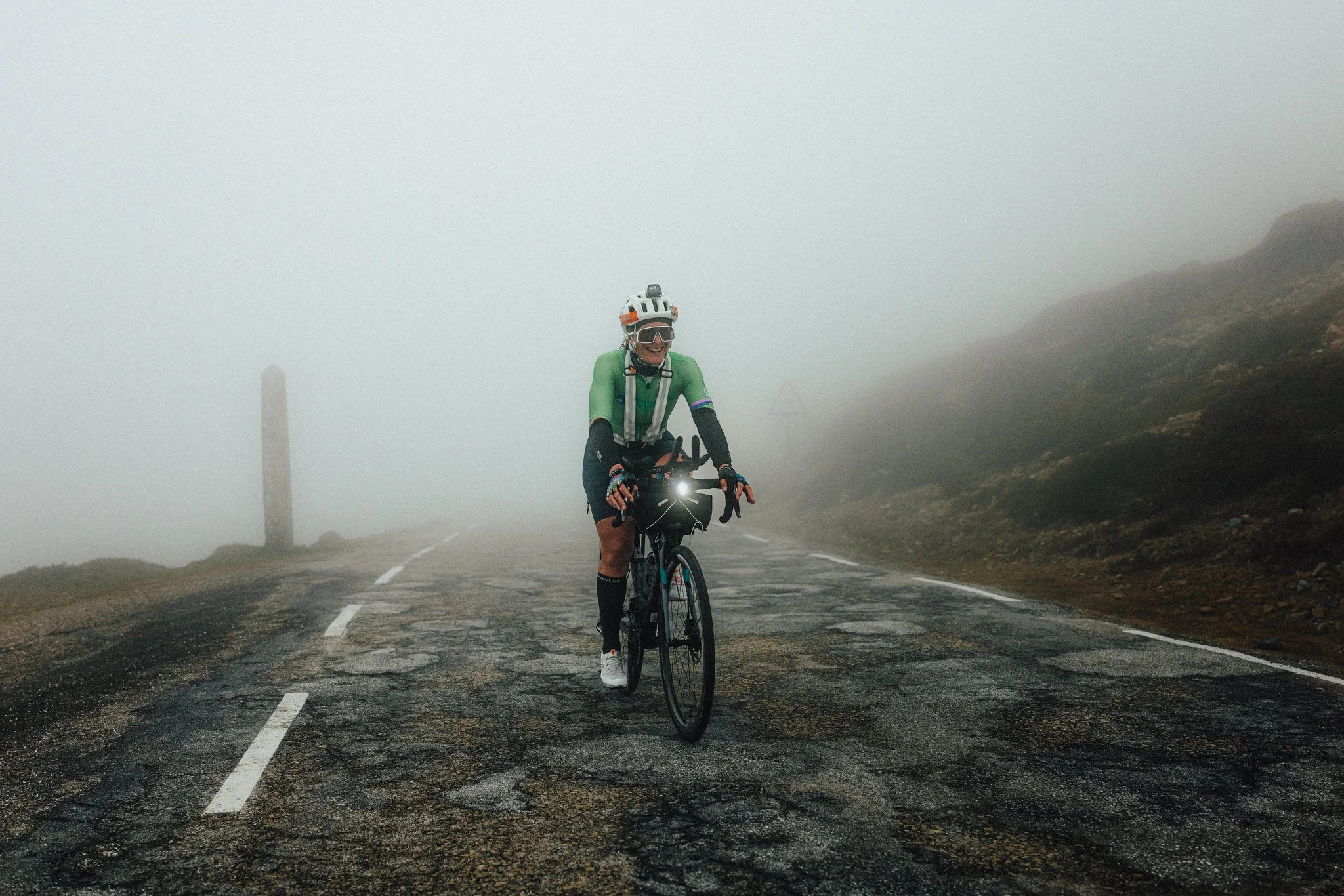 Un ciclista sonriente, con casco, gafas y maillot verde, pedalea por una carretera de montaña rota y con niebla.