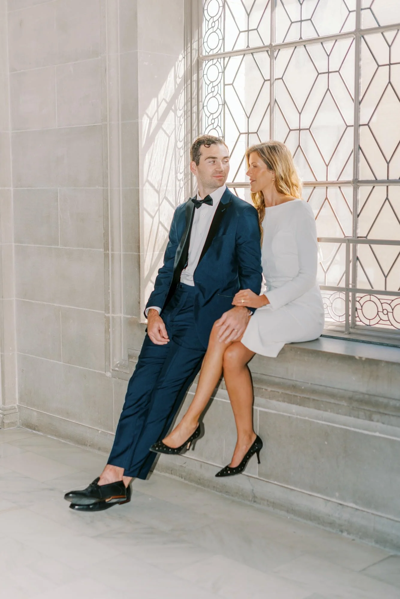 A man in a navy tuxedo and a woman in a white dress sitting on a stone windowsill in front of a decorative window, with sunlight casting shadows.