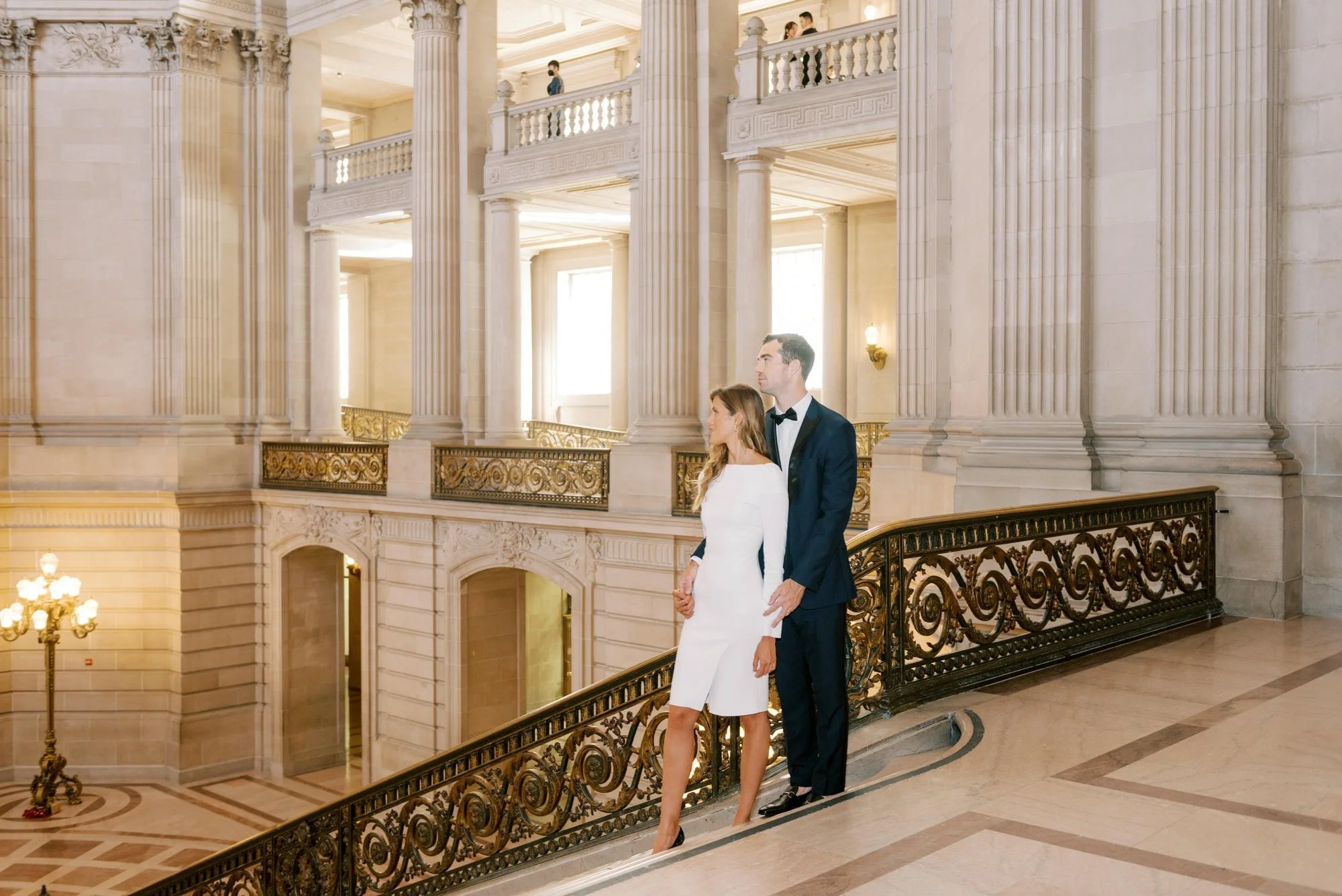 A man and a woman dressed in formal attire walking hand-in-hand on a marble balcony inside a grand, neoclassical building with tall columns and ornate gold railings.