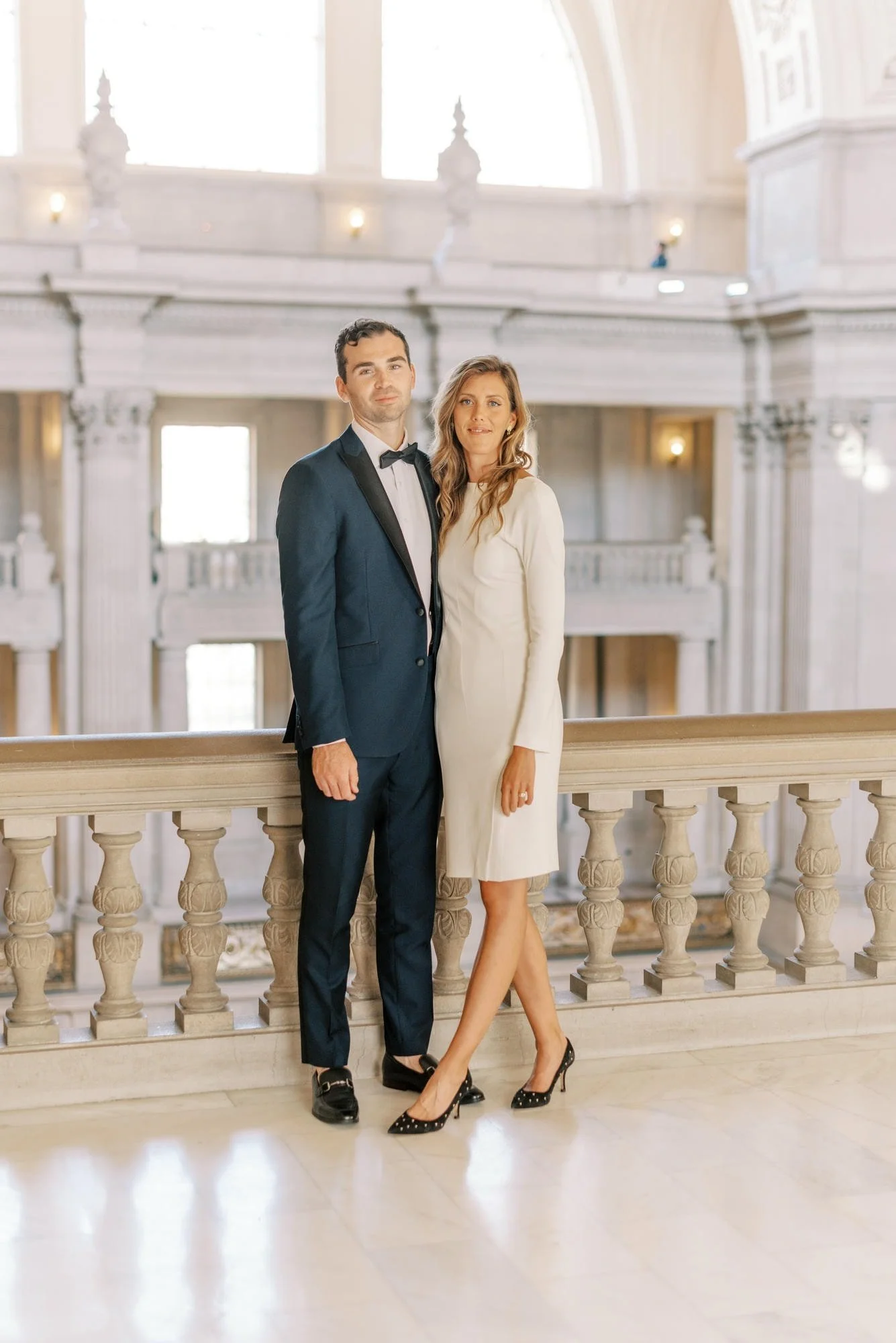 A couple dressed in formal attire standing on a balcony of a grand, ornate building with high ceilings and large windows.