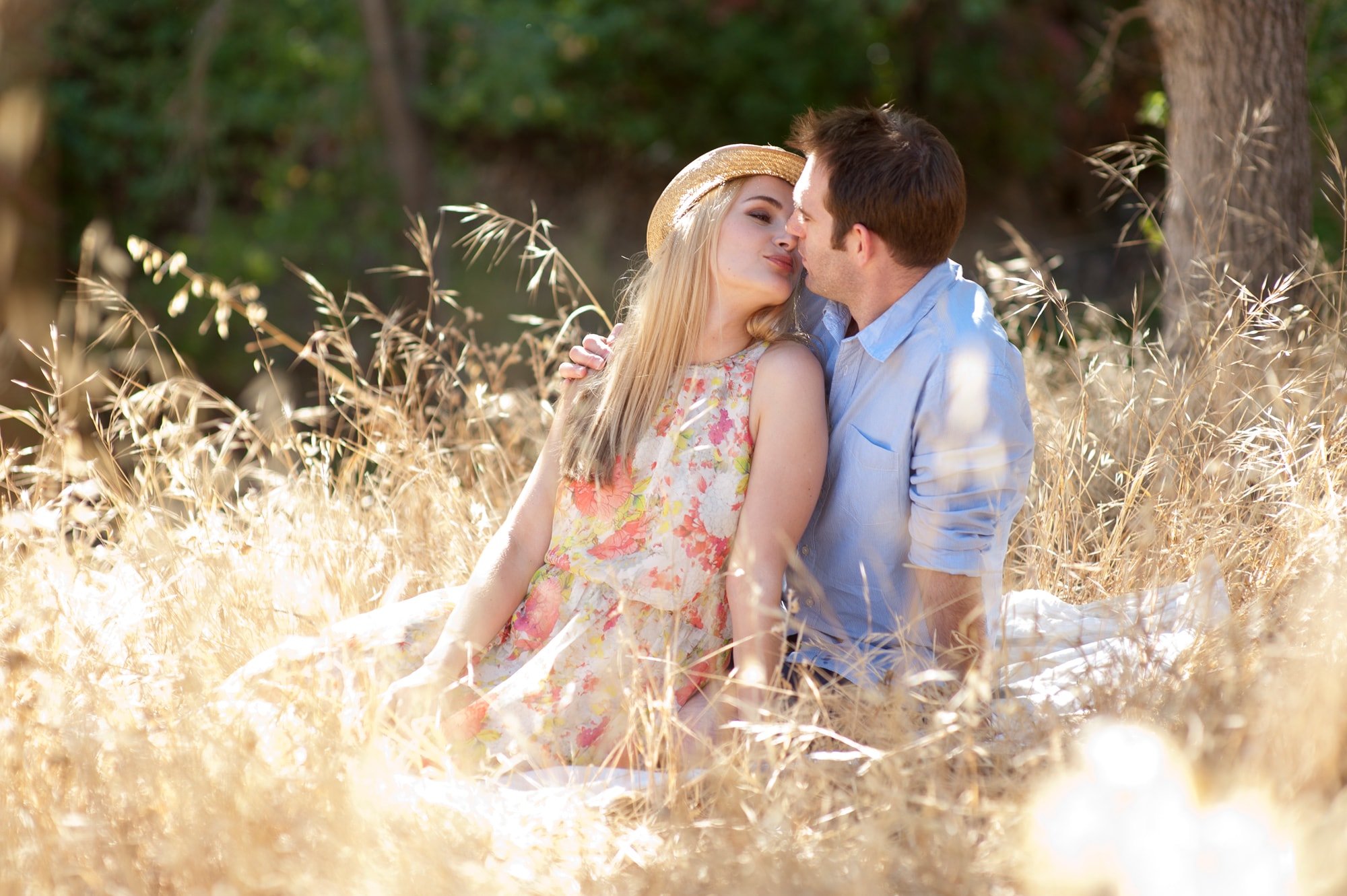 A couple sitting in a grassy field, sharing a tender moment with their faces close, surrounded by golden tall grass and trees in the background.