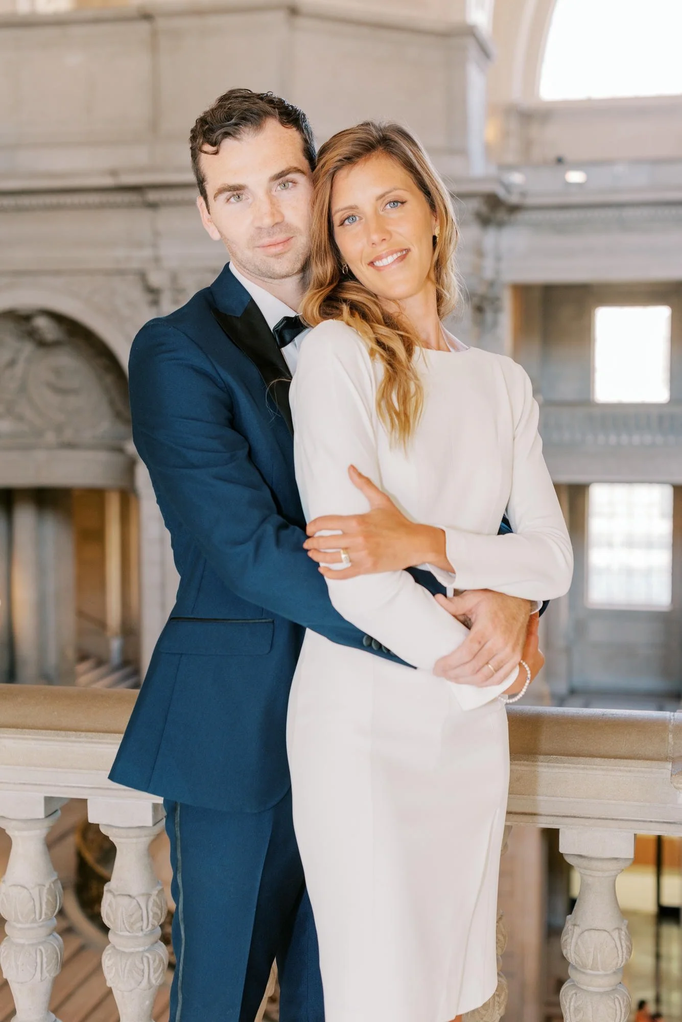 A couple dressed in formal attire standing close together in an elegant, historic building with large windows and ornate architecture.