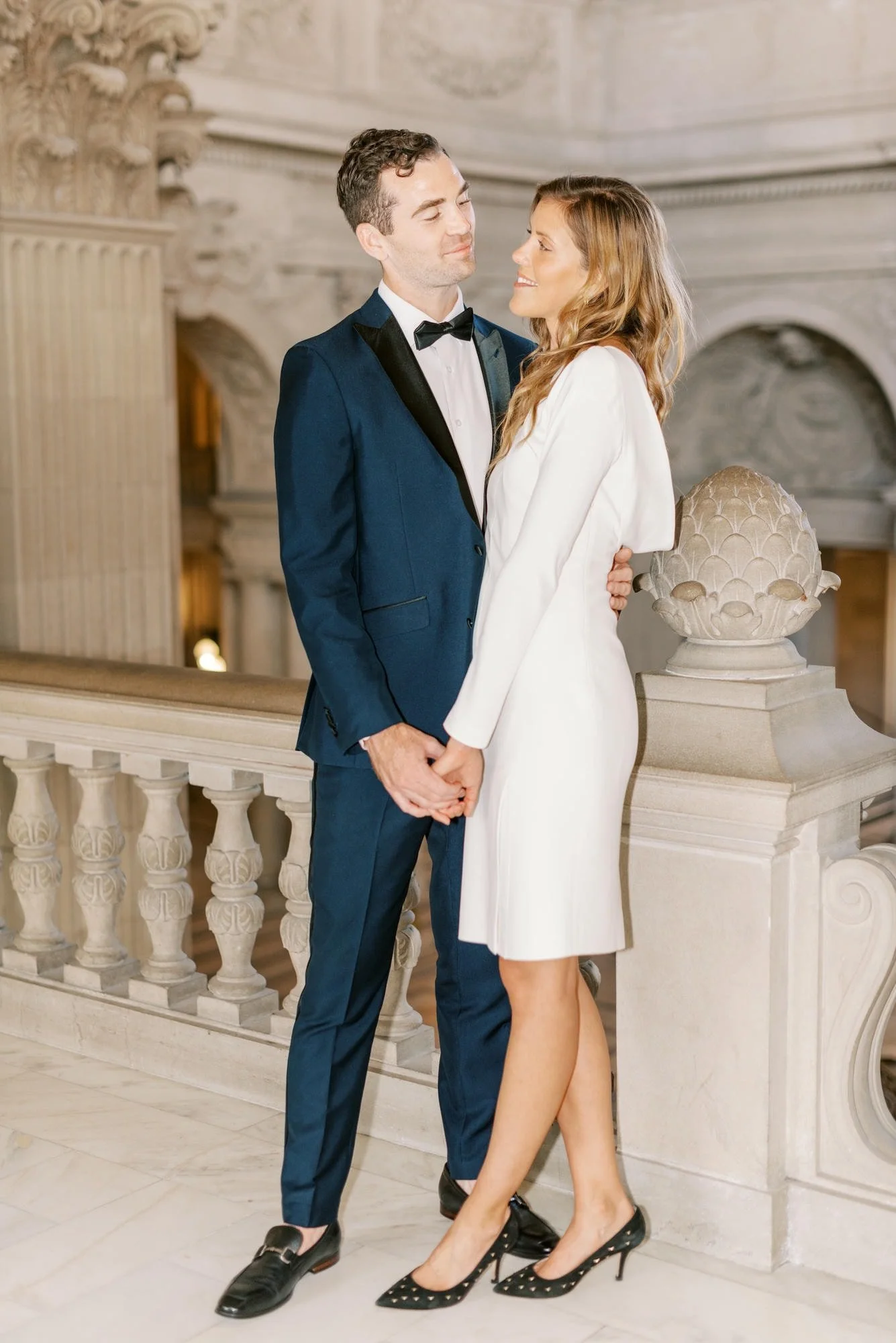 A man in a blue tuxedo and woman in a white dress holding hands and smiling at each other on a marble balcony in an ornate building.