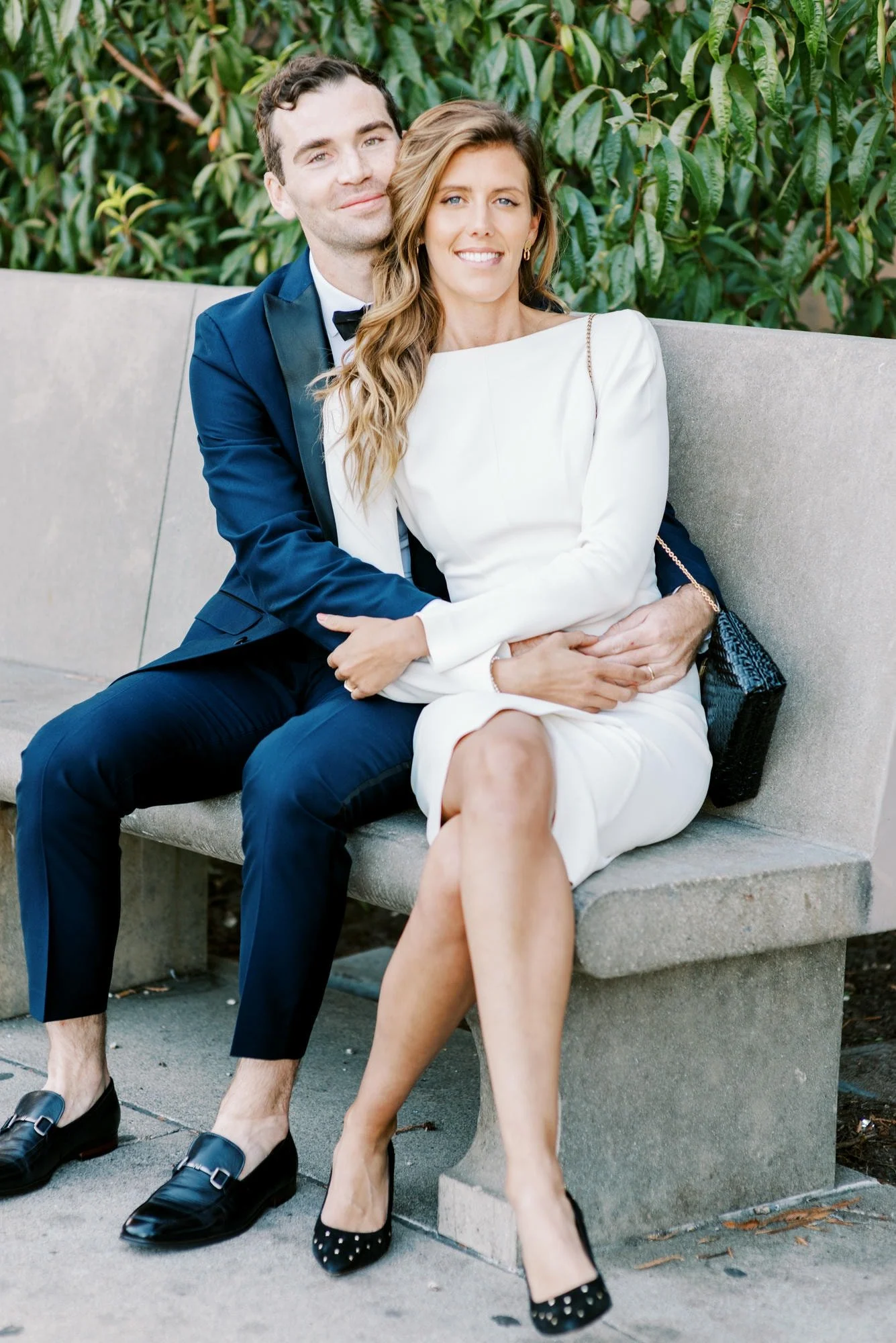 A couple dressed in formal attire sitting on a concrete bench outdoors, with green leafy background. The man wears a navy blue tuxedo with a black bow tie, and the woman wears a white dress and black heels. They are smiling, with the man's arms aroun