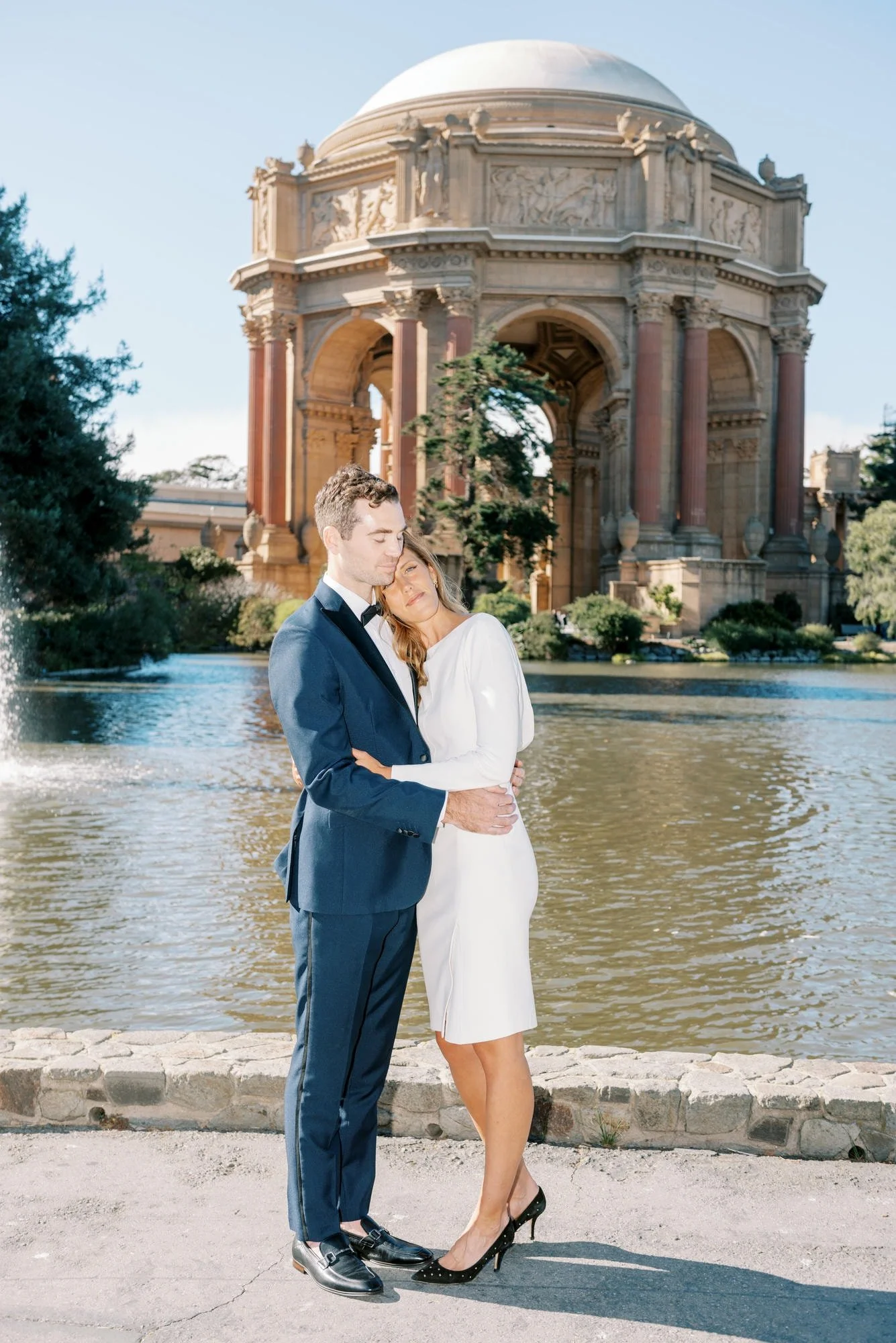 A couple embracing each other in front of a historic monument overlooking a body of water. The man is wearing a blue suit and the woman is in a white dress and black high heels. The scene is outdoors on a sunny day.