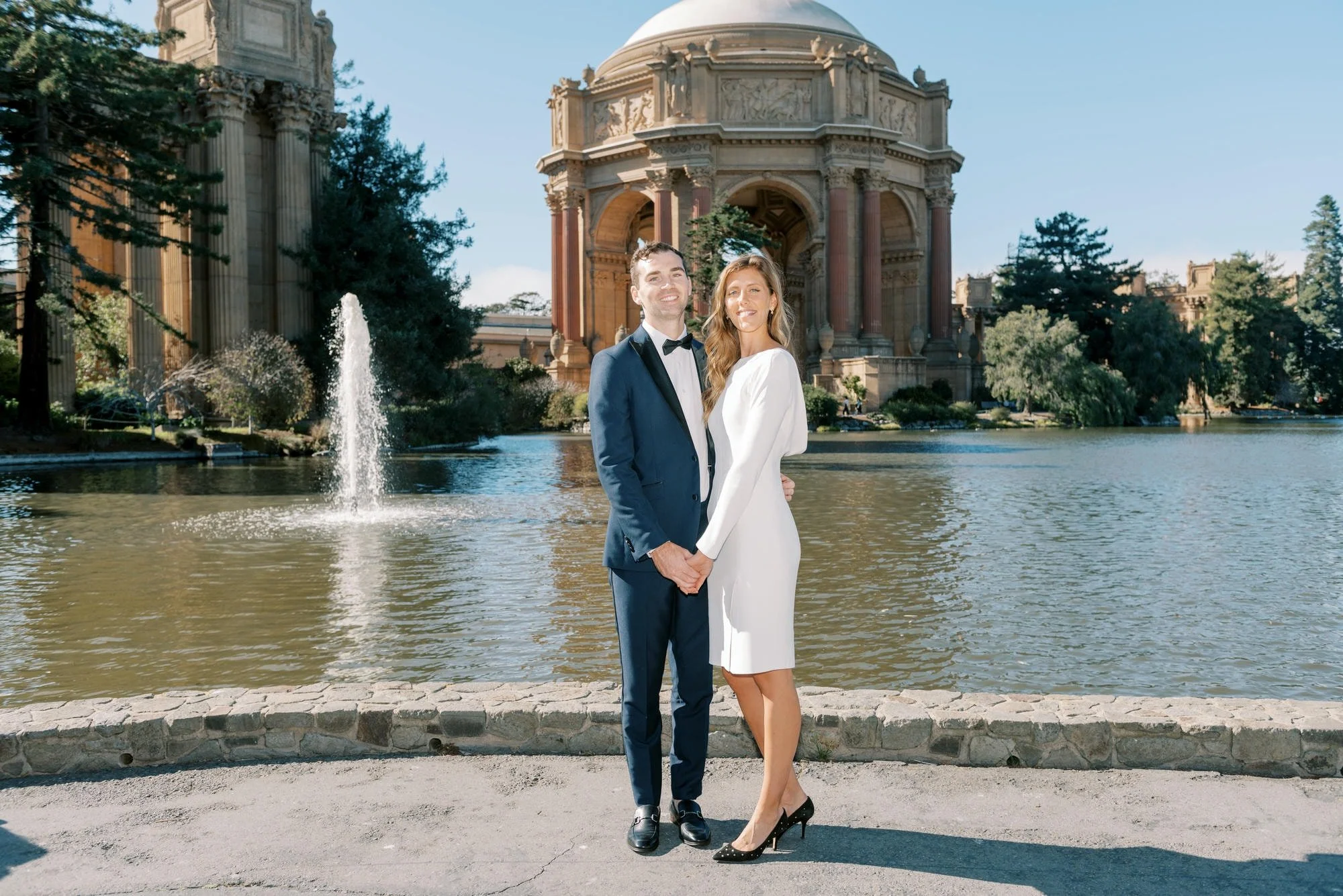 A couple in wedding attire stands hand in hand by a pond with a historic temple in the background, on a sunny day.