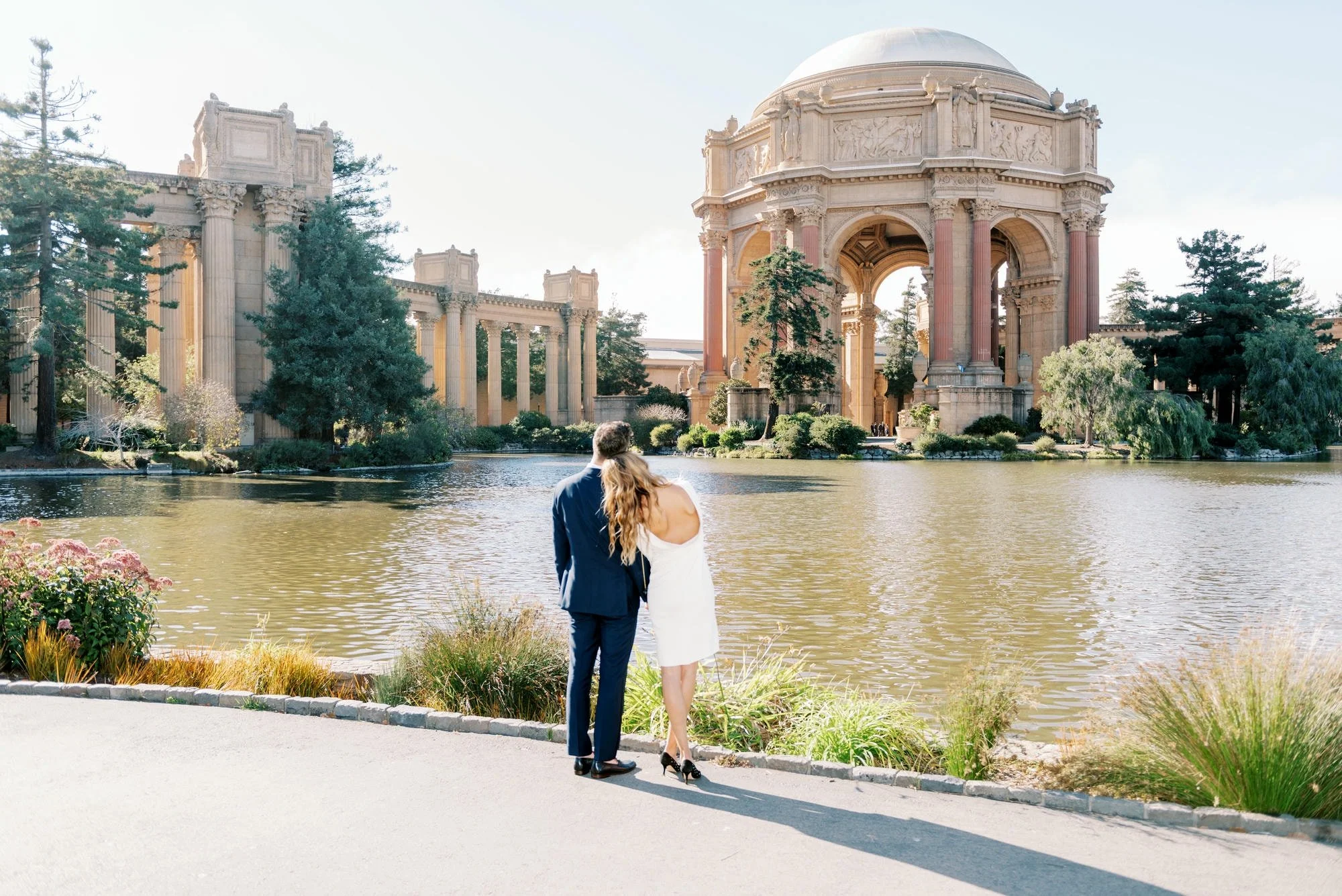 Couple standing by a pond with a historic architectural backdrop, including tall columns and a domed structure, on a sunny day.