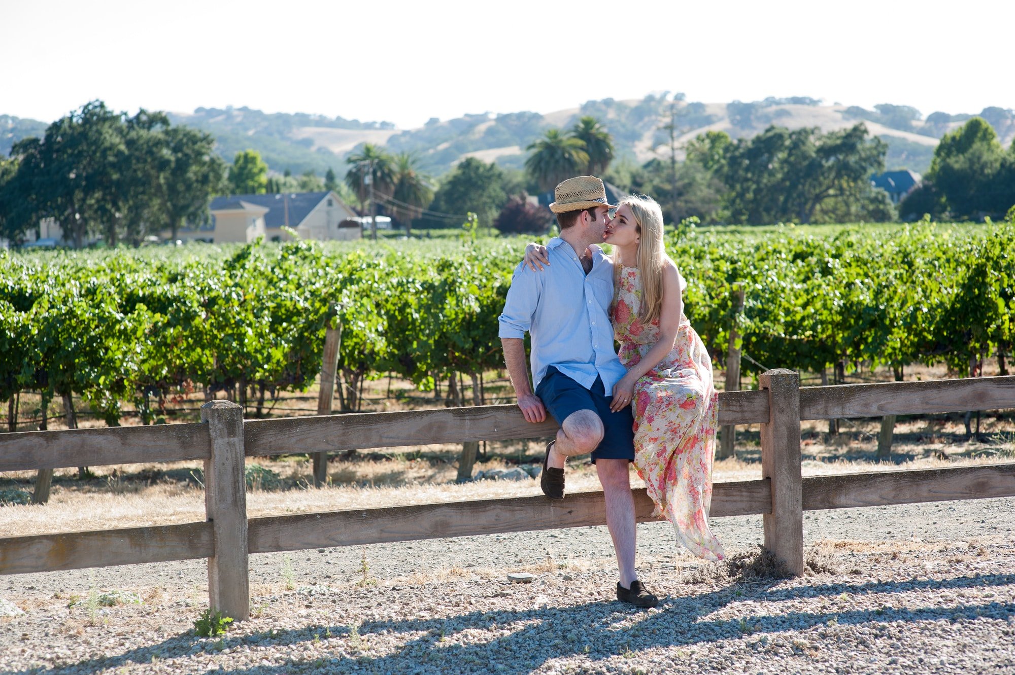 A young couple kissing on a wooden fence in a vineyard on a sunny day with rolling hills and trees in the background.