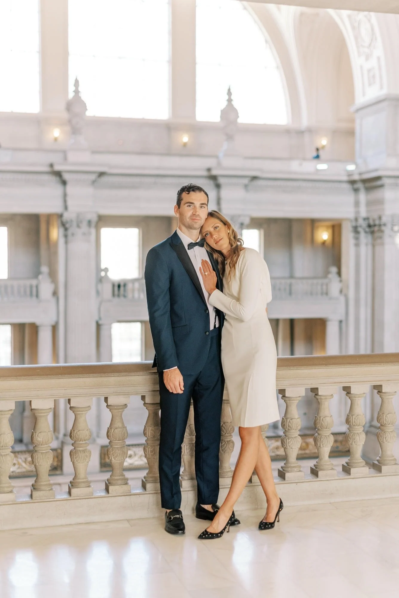 A man in a tuxedo and a woman in a white dress standing close together in an elegant, historic building with large windows and ornate architecture.