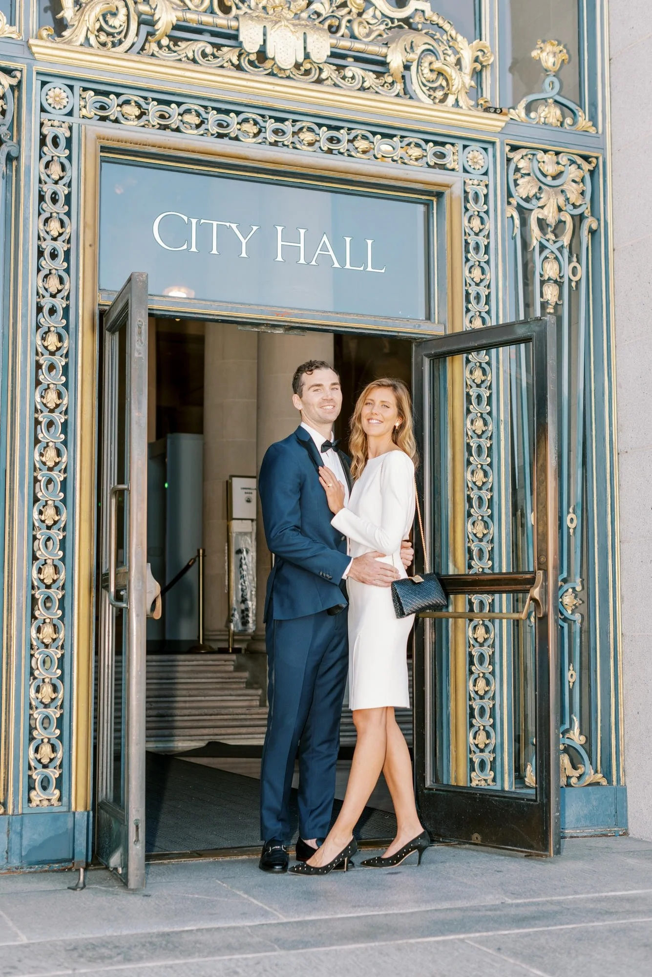 A couple dressed in formal attire standing in front of the entrance to a city hall building, smiling and posing for the photo.