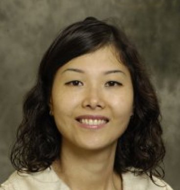 A woman with shoulder-length curly black hair, light skin, and a friendly smile, wearing a beige top, against a gray background.