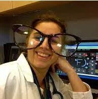 A woman in a white lab coat smiling in a medical or laboratory setting, wearing large glasses, with a microscope in the background.