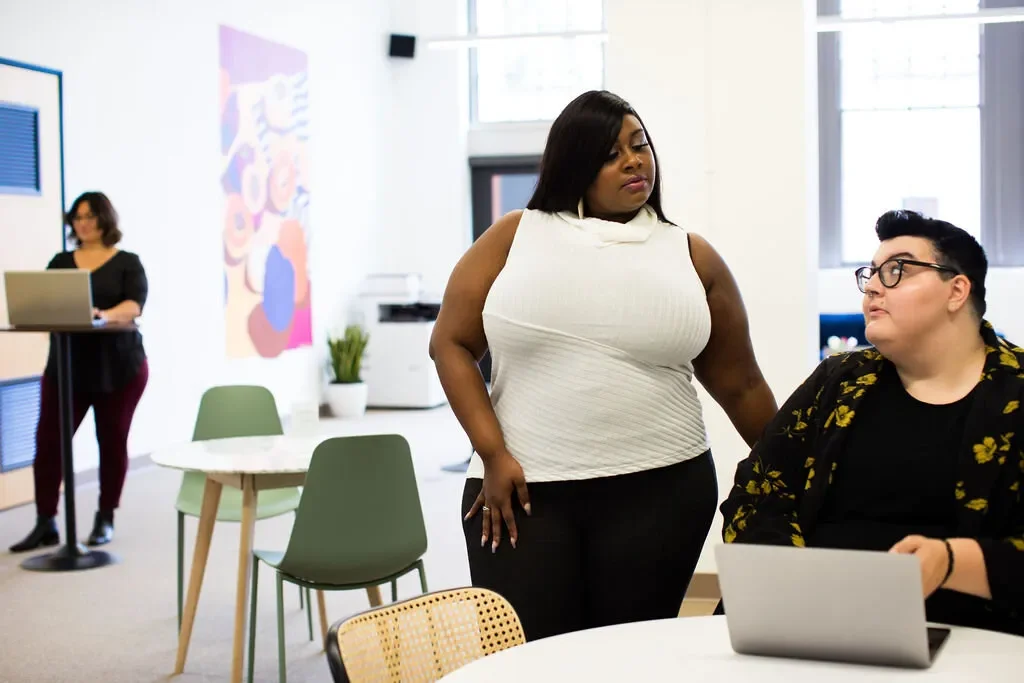 Two plus sized people work together looking at something on a computer. One is a femme Black person, wiht long straight hair, and a white shirt. The other has light skin, dark short hair, and is wearing glasses.