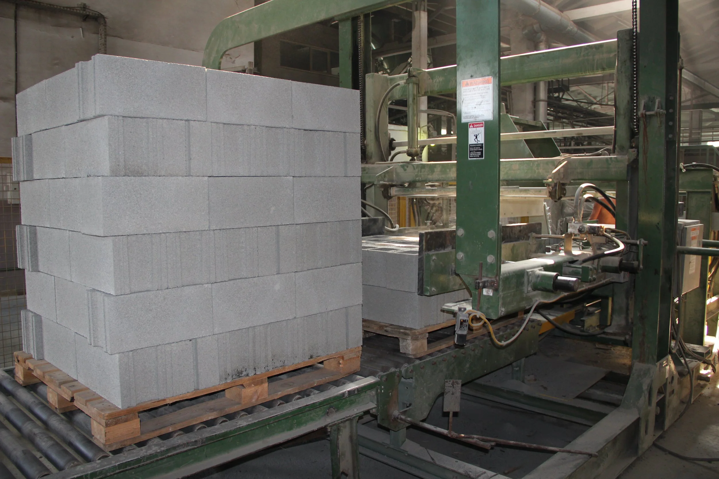 Cinder blocks stacked on a pallet beside a green brick-making machine in an industrial setting.