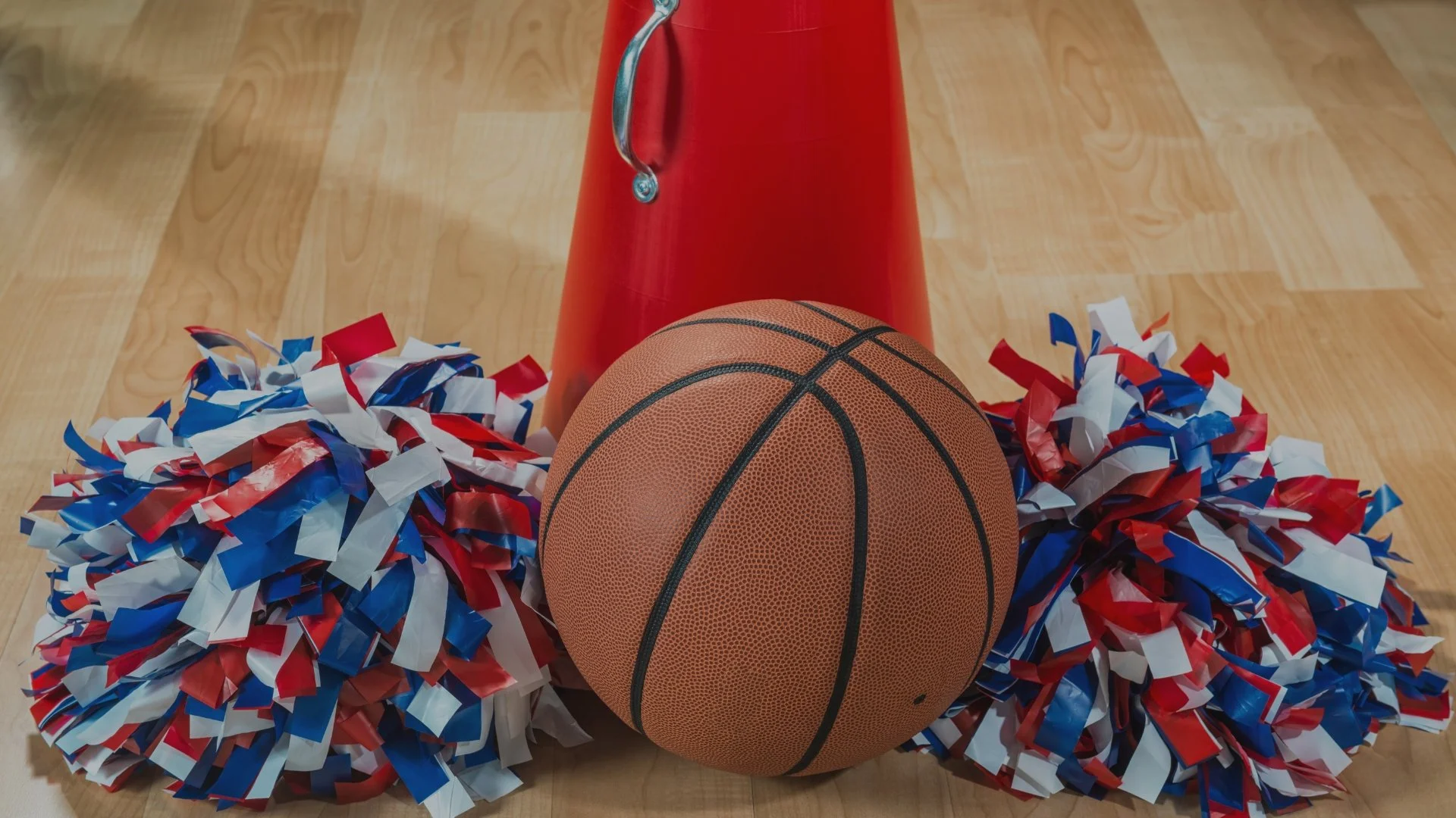 Basketball, red megaphone, and red, white, and blue pom-poms on a wooden gym floor.