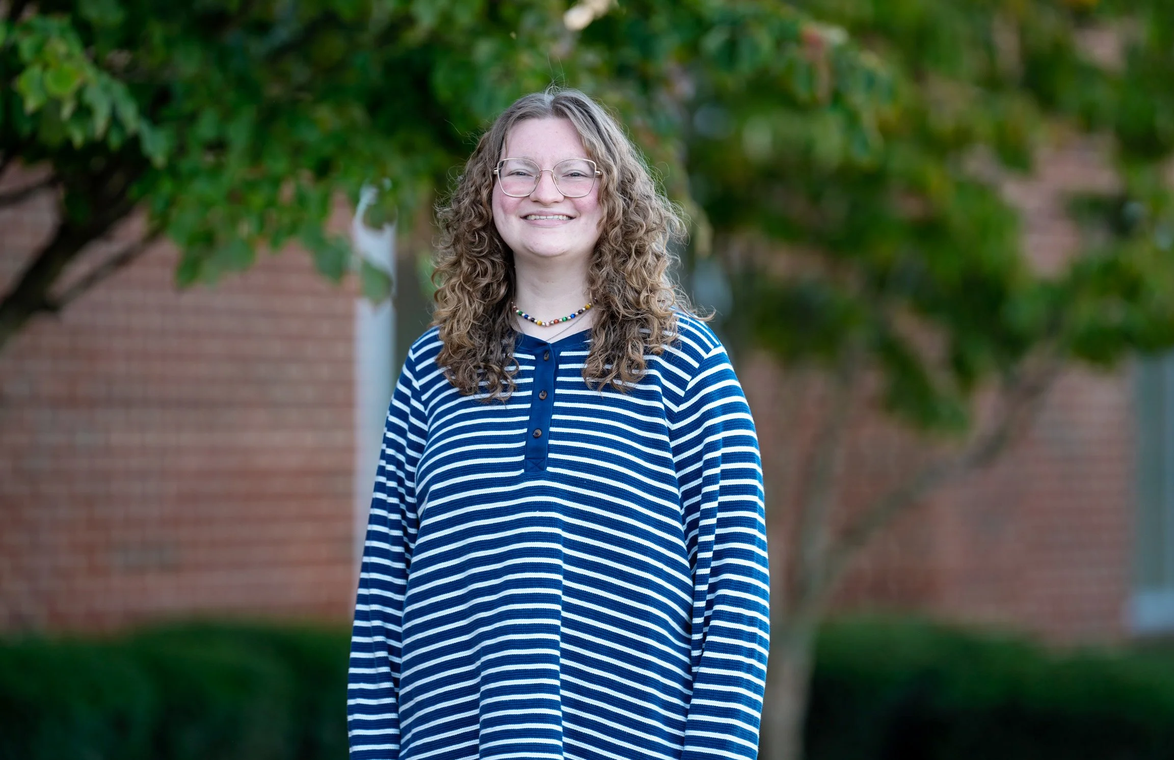 A young woman with curly hair, glasses, and a beaded necklace standing outdoors near trees and a brick building, smiling at the camera.