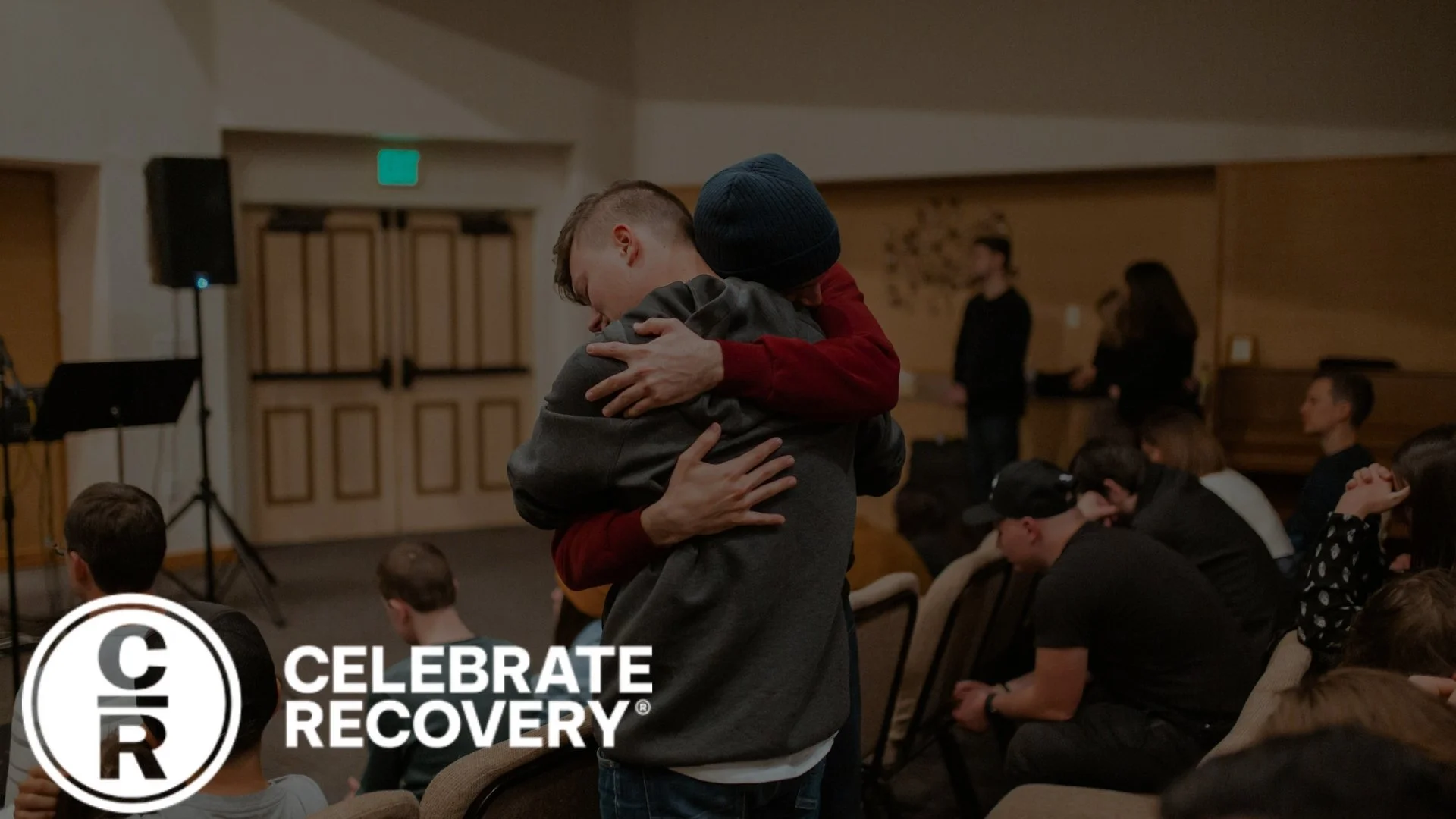 Two men hugging at an indoor event with seated audience members in the background and a logo reading 'Celebrate Recovery' in the lower left corner.