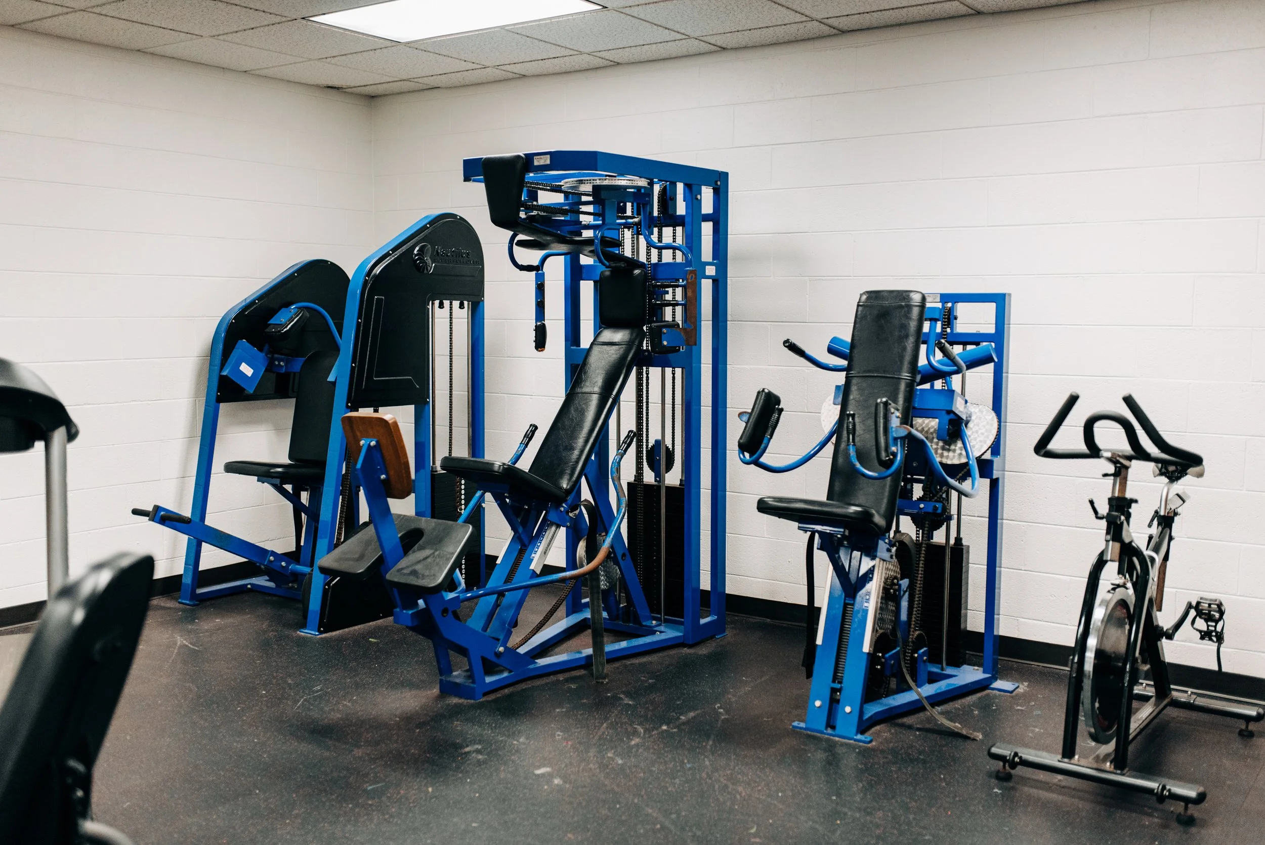 Gym room with various exercise equipment, including a stationary bike, resistance machines, and a weight bench, against white cinder block walls.