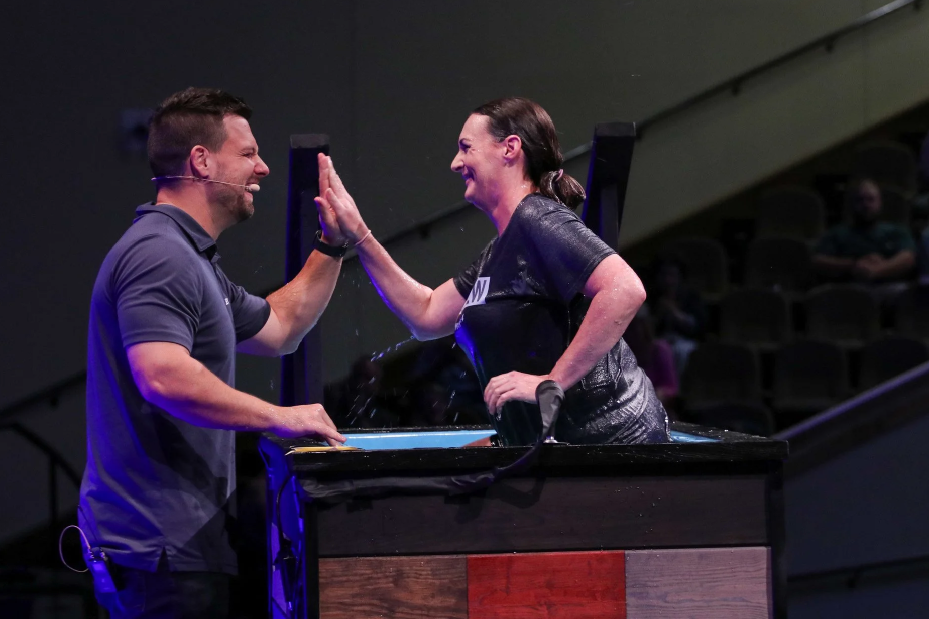 A man and a woman in water baptism are smiling and high-fiving each other inside a baptismal pool. The woman is wearing a dark shirt, and the man is wearing a gray shirt. The background shows an auditorium with dark seating.