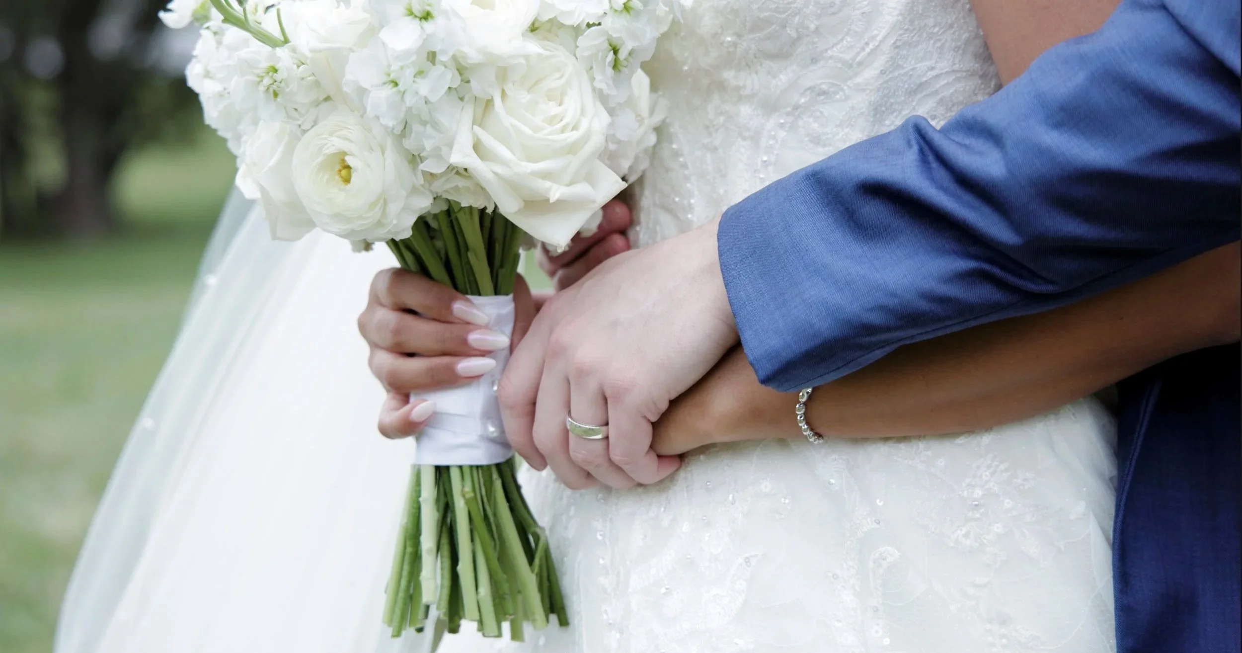 Close-up of a bride and groom holding hands, with the bride holding a bouquet of white roses and greenery, outdoors with a blurred green background.